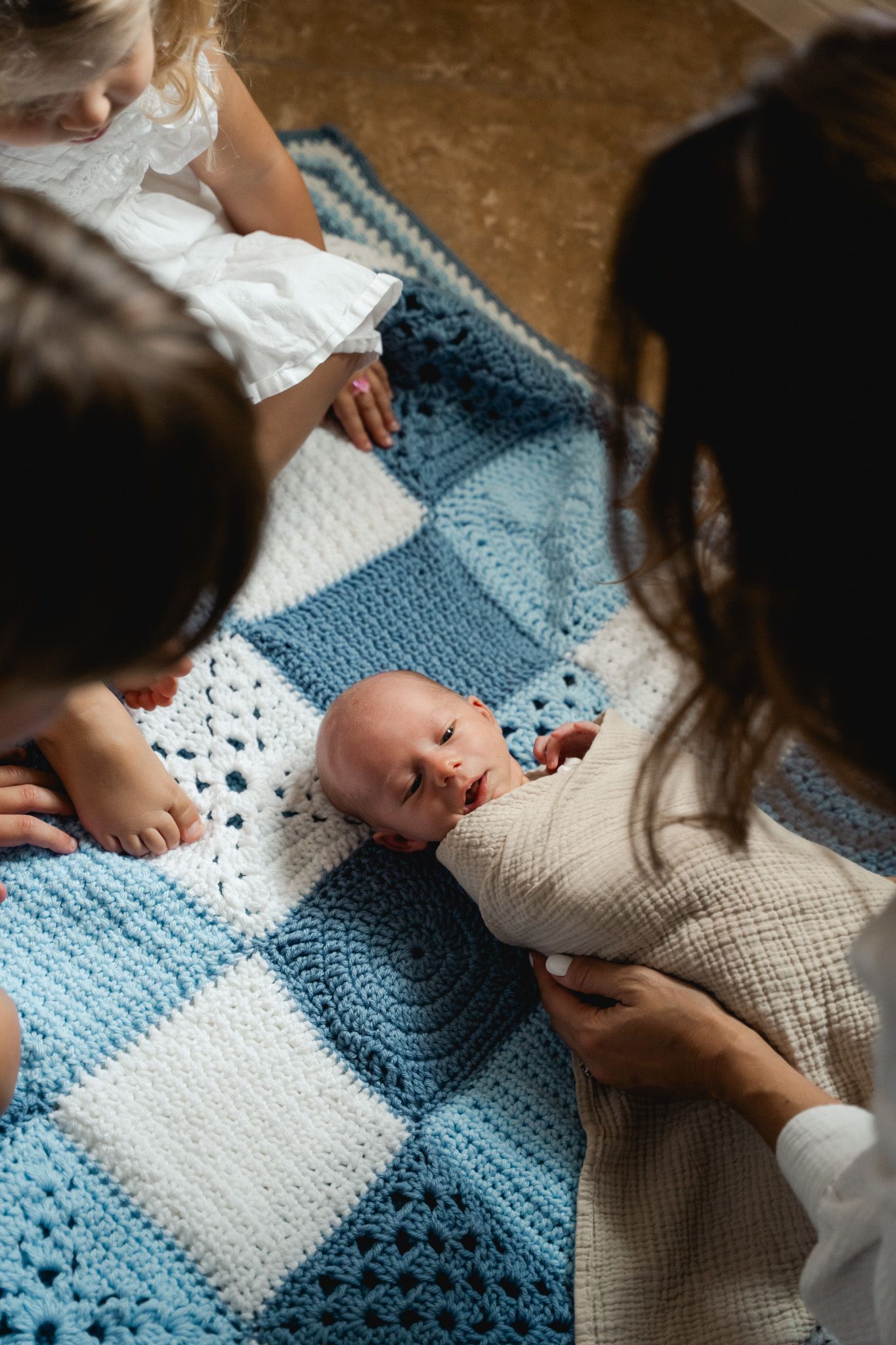 Family gathered around a swaddled newborn lying on a blue and white crocheted patchwork blanket.