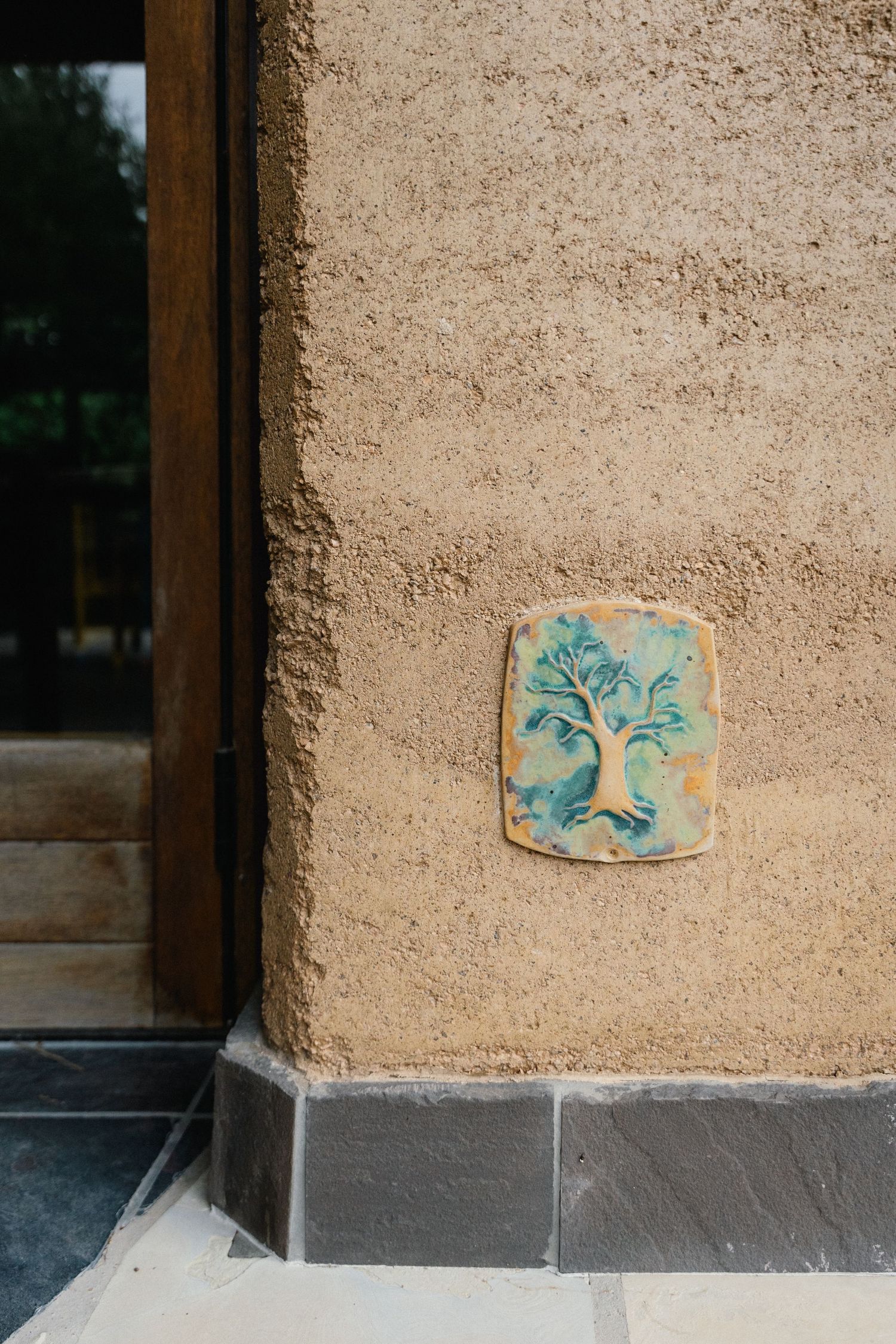 Decorative ceramic tile plaque featuring a blue-green tree design mounted on a textured beige stucco wall.