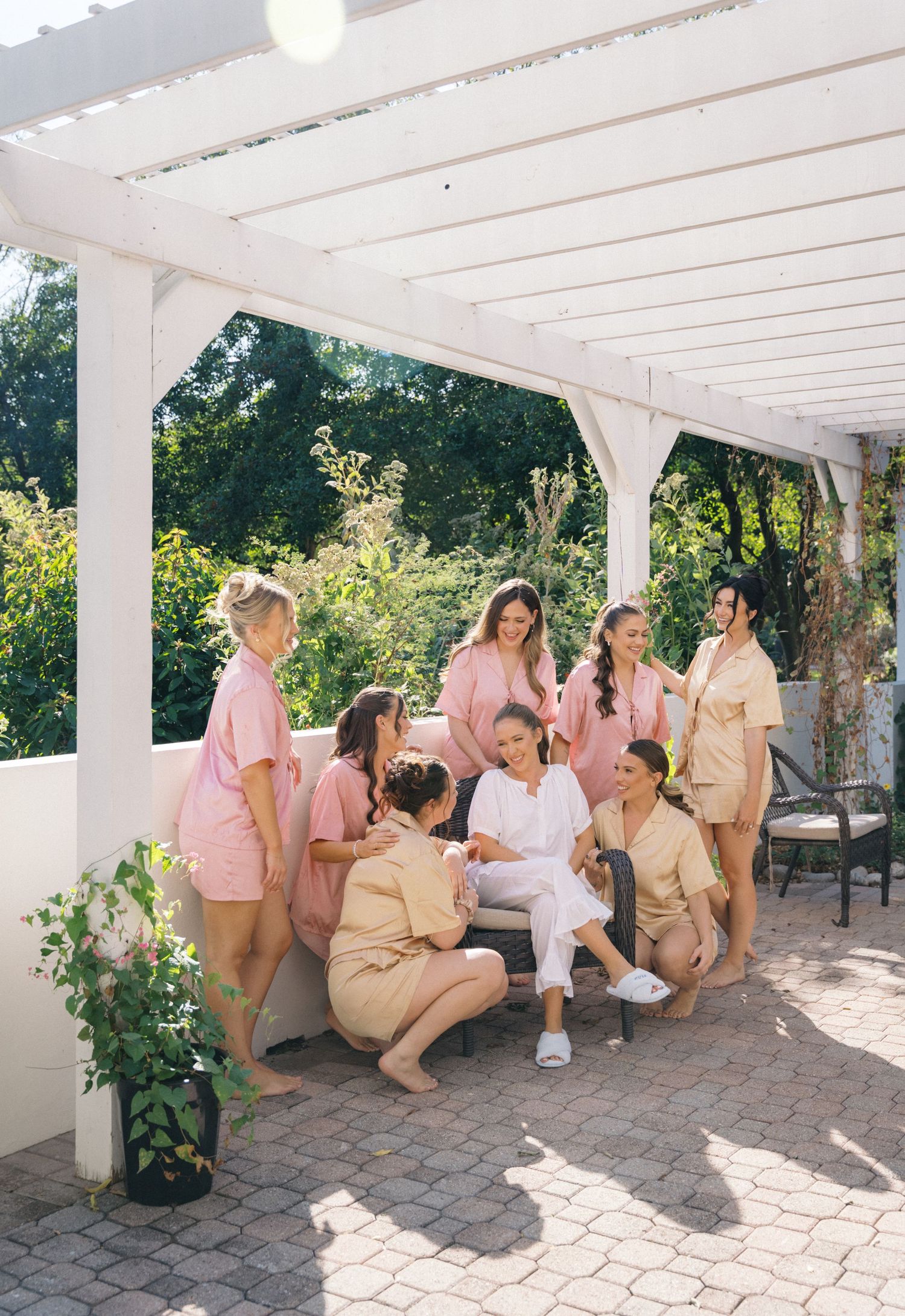 Bride in white relaxes with bridesmaids in pink and tan matching sets under white pergola on sunny patio.