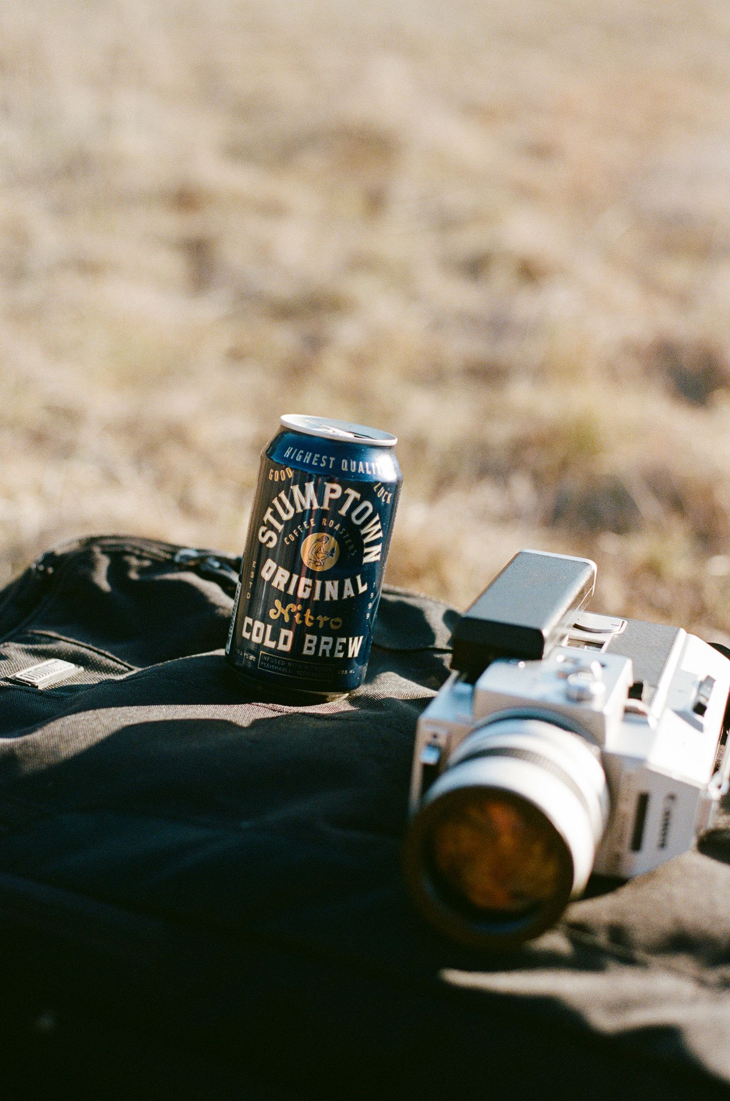 Stumptown Cold Brew coffee can beside a film camera on a dark jacket outdoors.