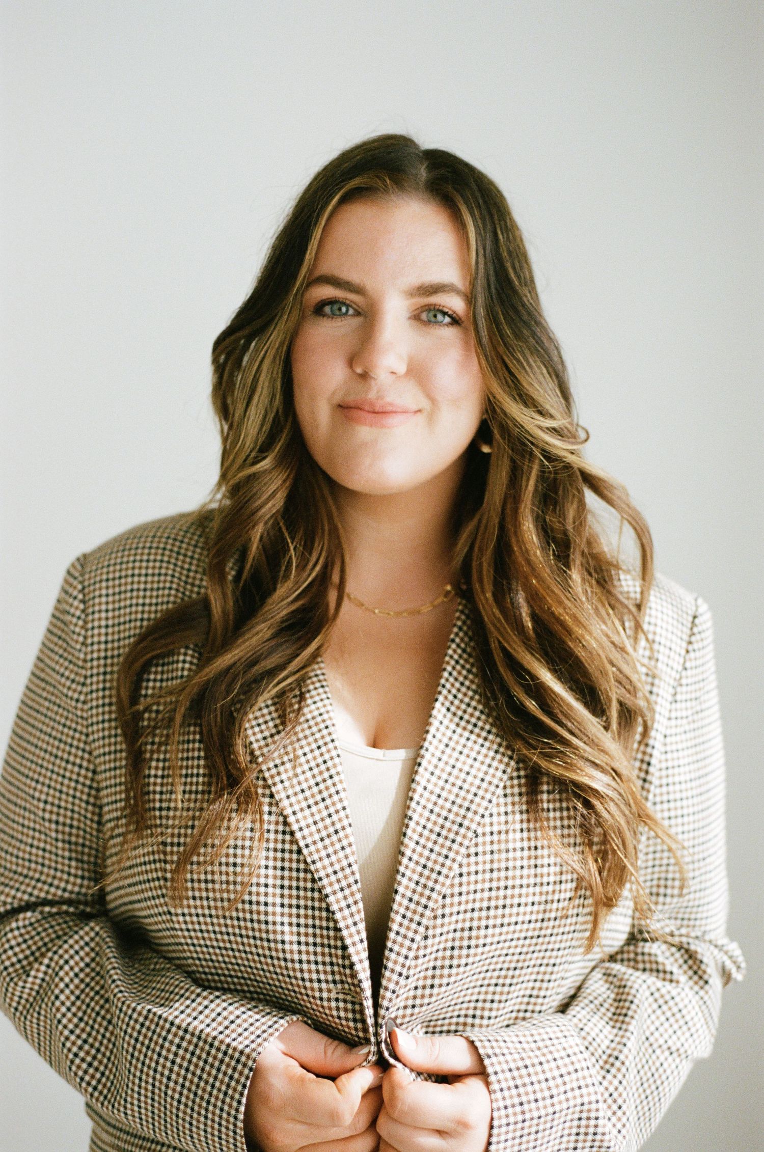 Smiling brunette woman in houndstooth blazer with long wavy hair against a light gray background.