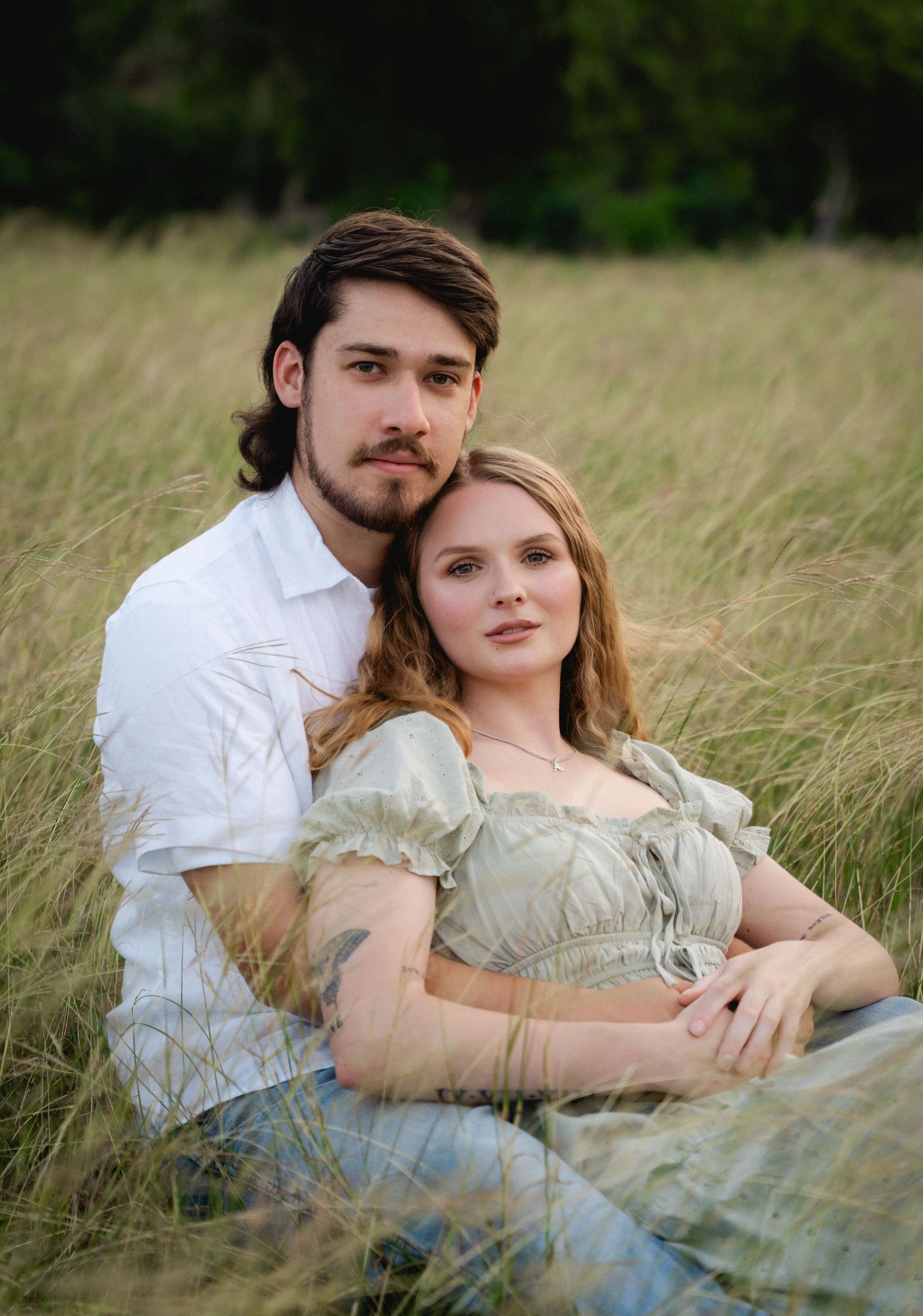 A couple sits together in a golden grass field, the woman in a sage dress cradled by her partner in a white shirt.