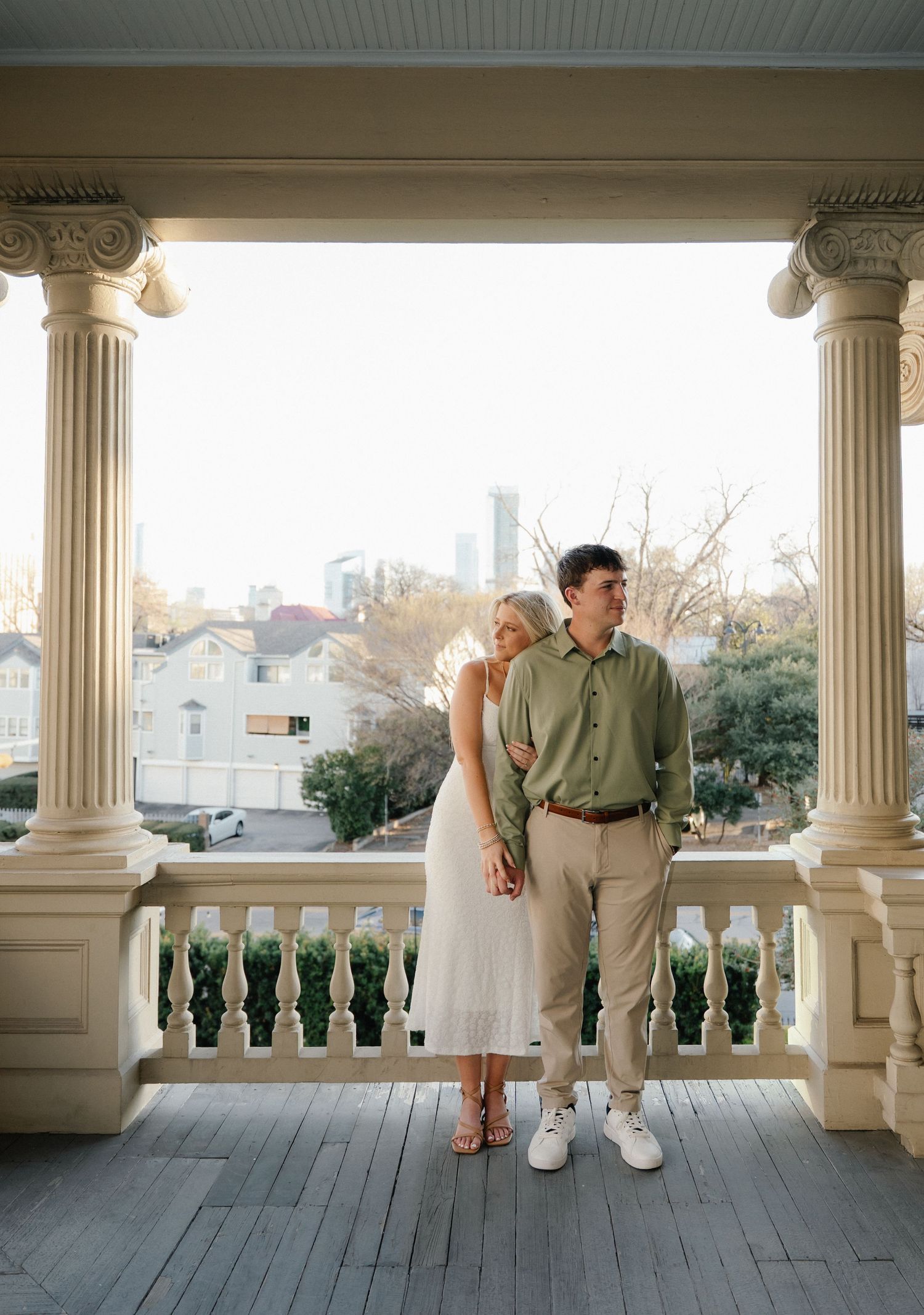 Couple posing on elegant white columned porch with city skyline backdrop, woman in white dress, man in green shirt.