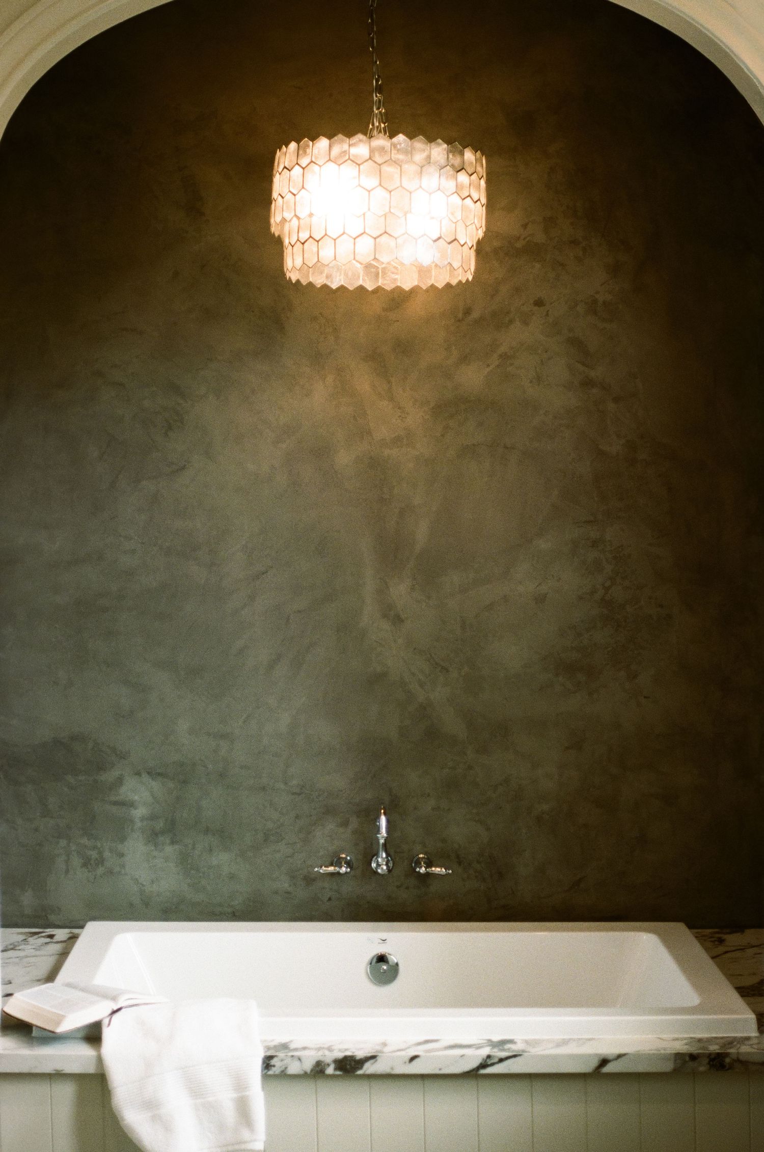 Elegant bathroom with freestanding white tub, marble surround, and capiz shell chandelier on dark wall.