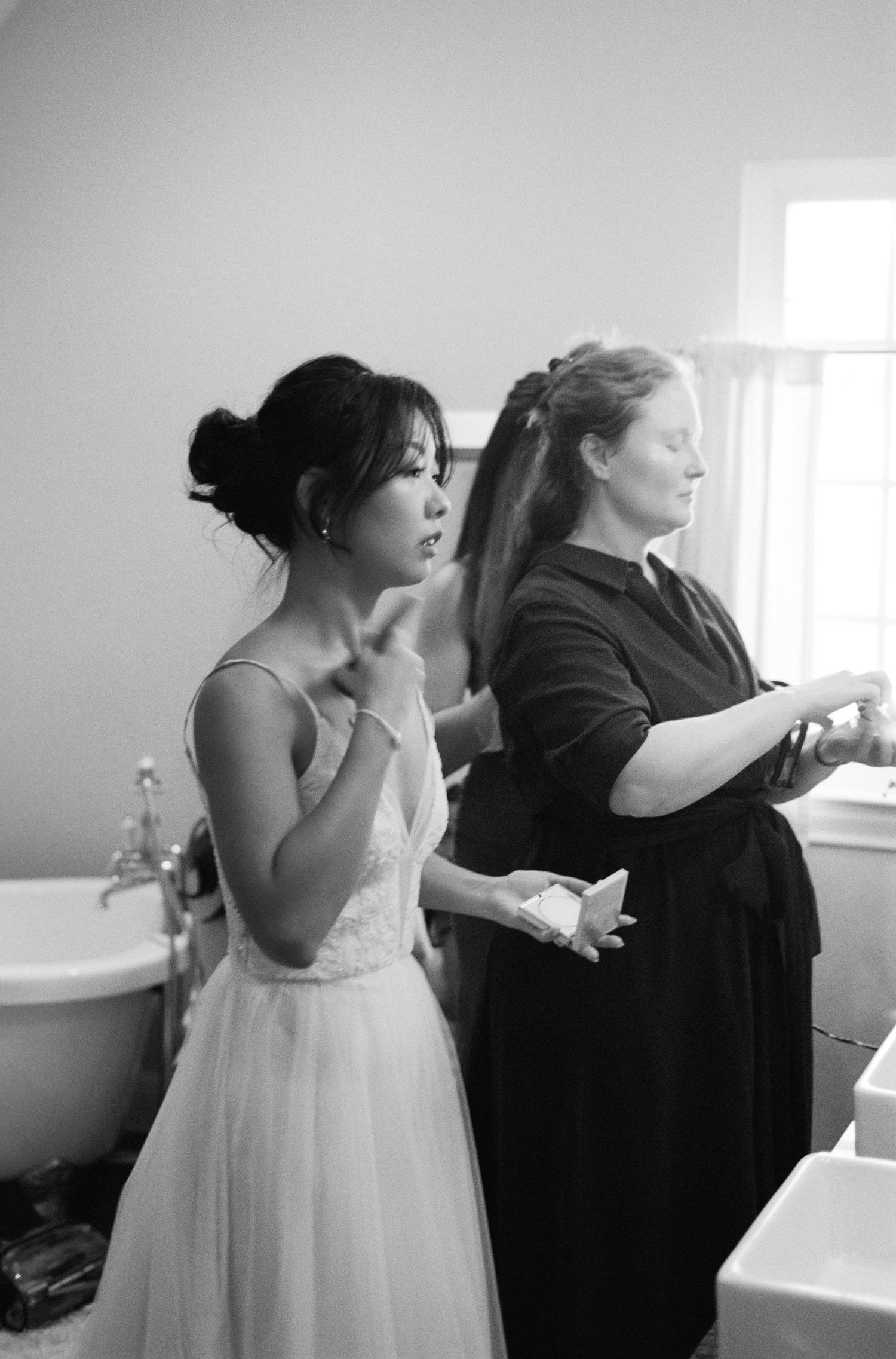 Bride in white gown gets hair styled by assistant in black and white bathroom photo.