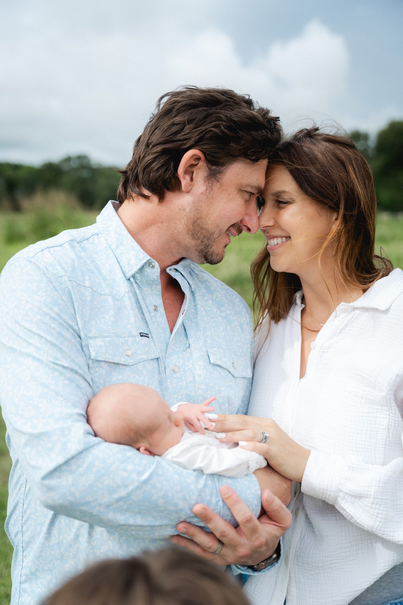 Smiling new parents touch foreheads while holding their newborn baby together in an outdoor field.