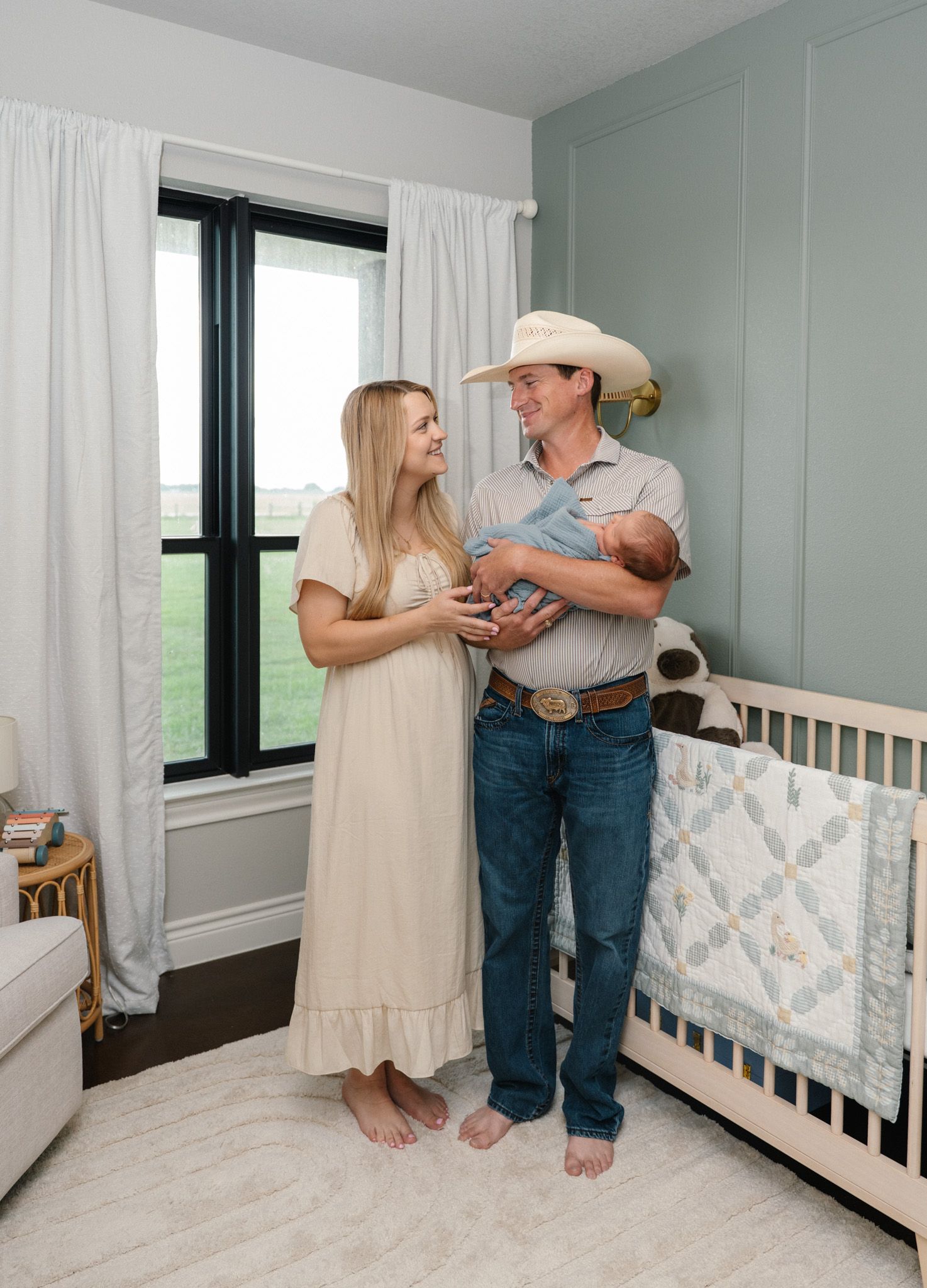 New parents in a nursery, father in cowboy hat holding newborn baby, mother in cream dress smiling.