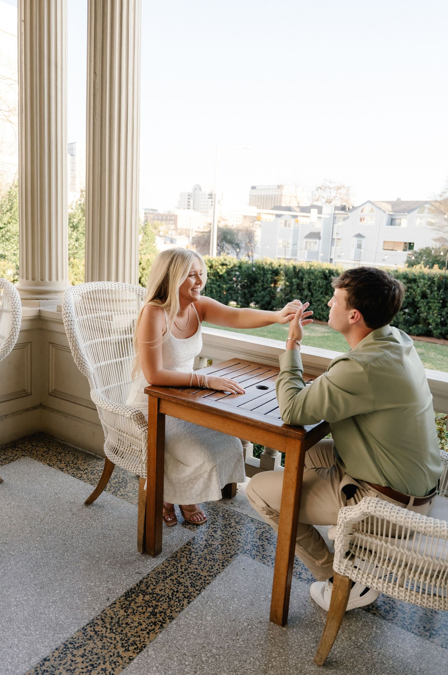Smiling couple holding hands across a wooden table on a sunlit columned porch with city views.