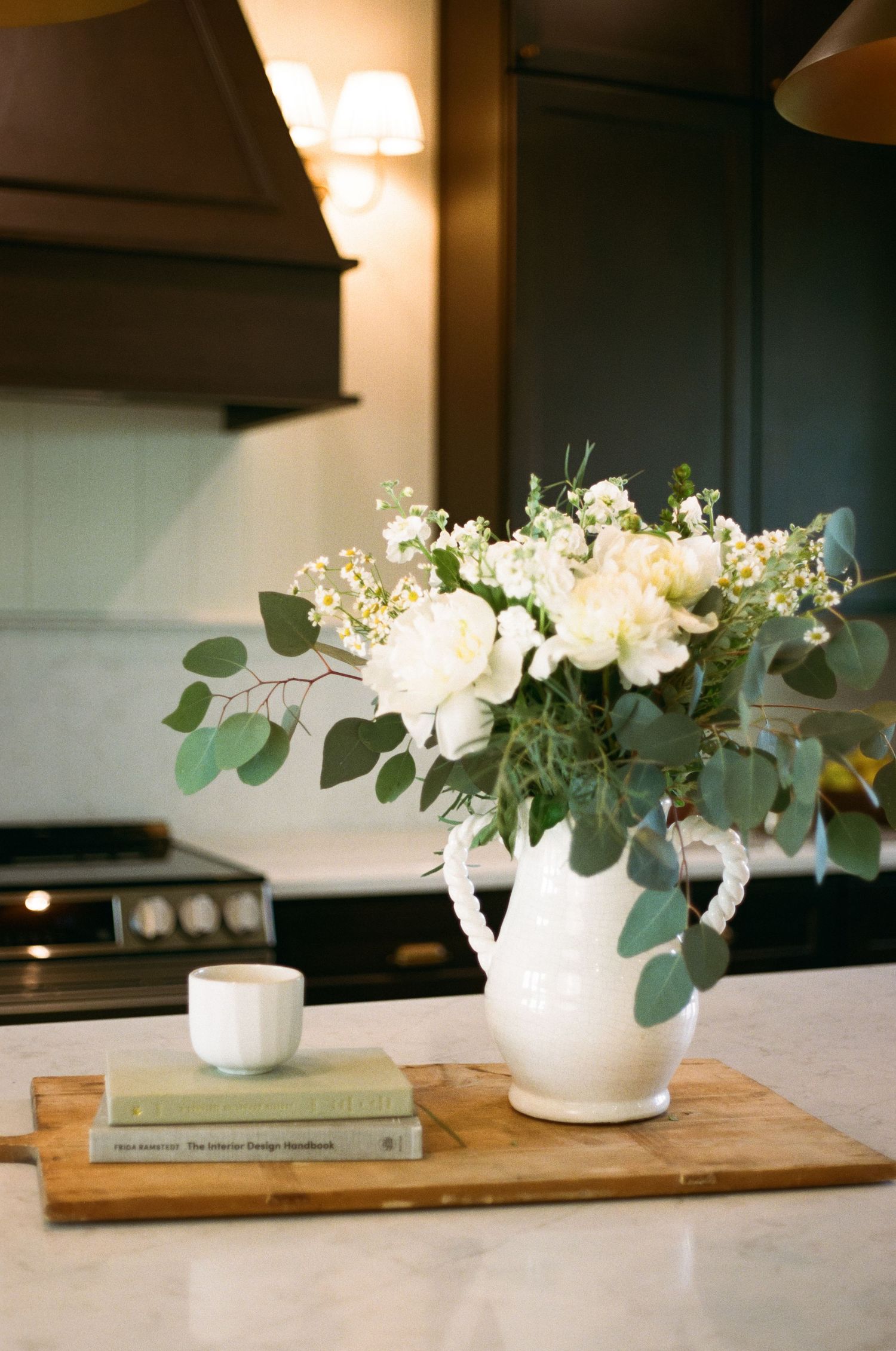 White pitcher with fresh eucalyptus and white roses on marble kitchen island beside stacked books and small white cup.