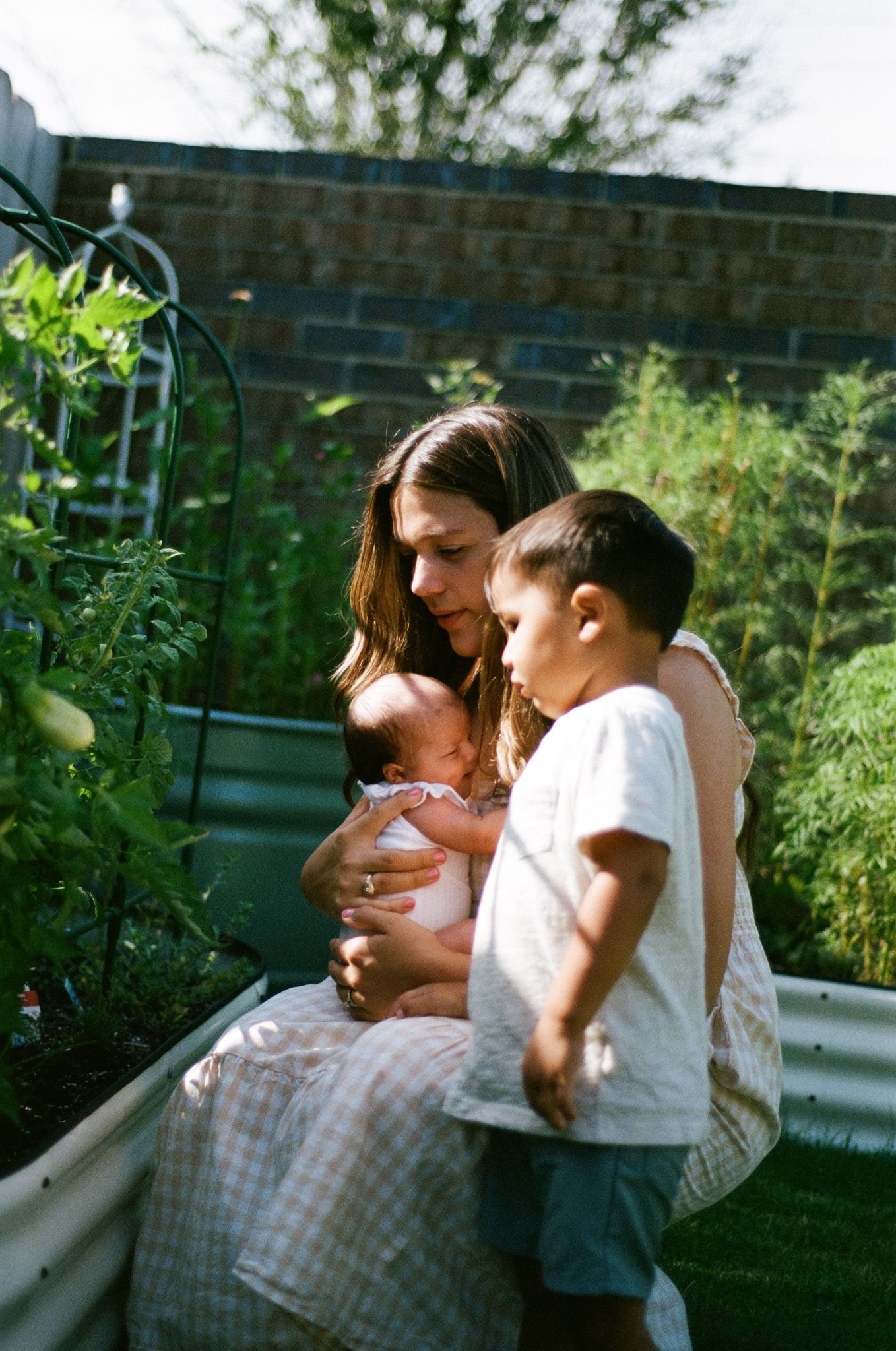A mother holds her newborn while her young son looks on in a lush garden with raised beds.