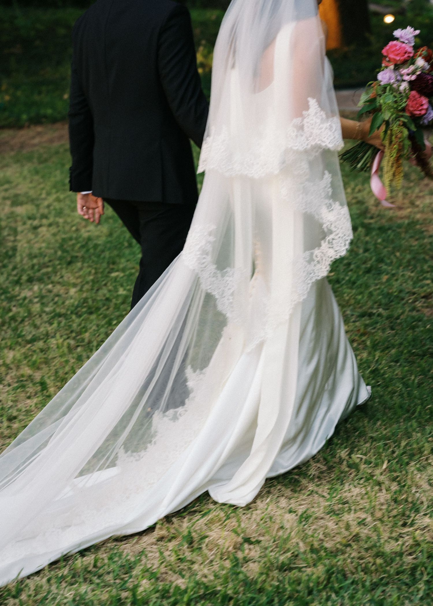 Bride in elegant lace-trimmed cathedral veil and white gown walks with groom in black suit on green grass.
