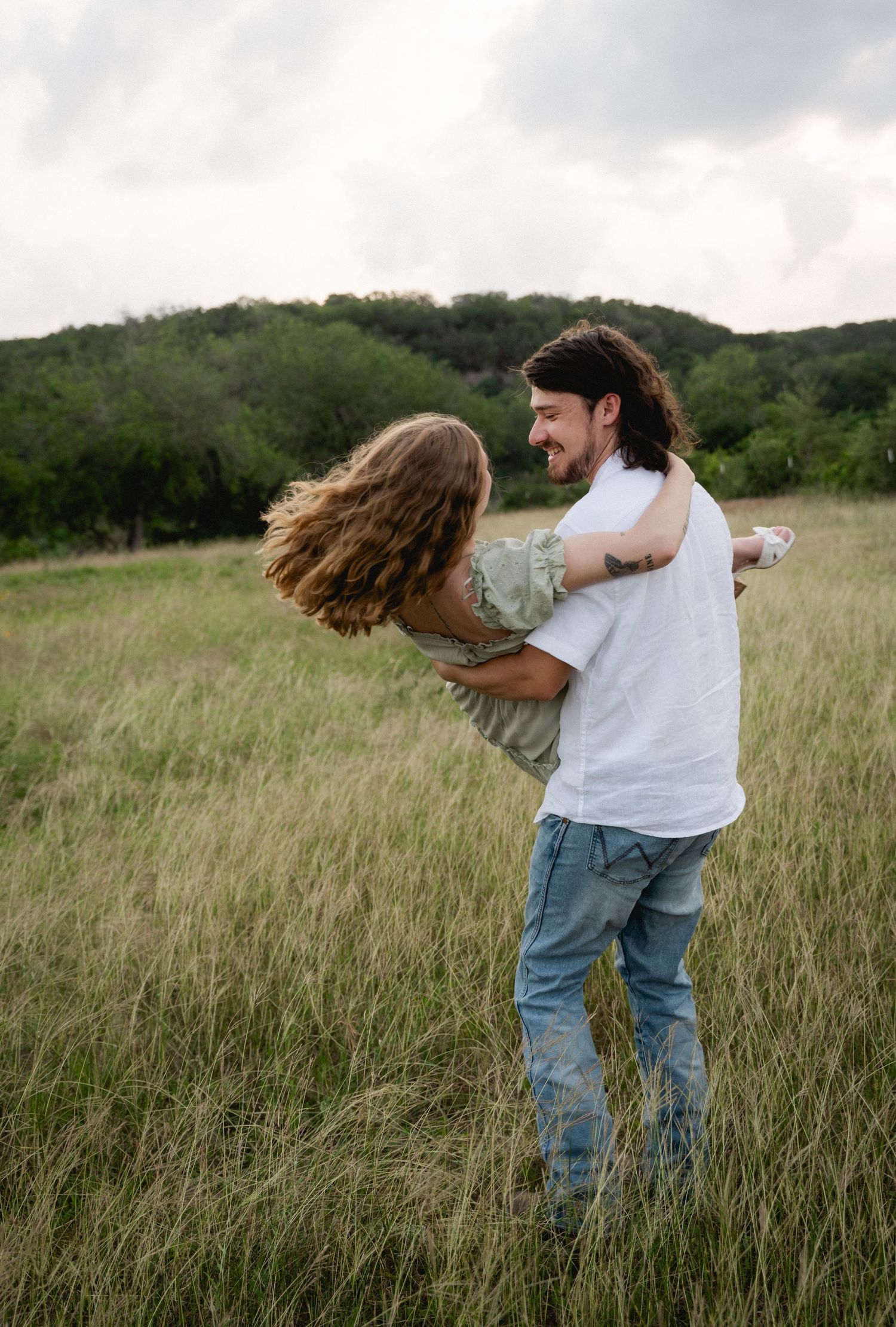 A man joyfully spins a woman in a grassy field with rolling green hills and cloudy skies in the background.