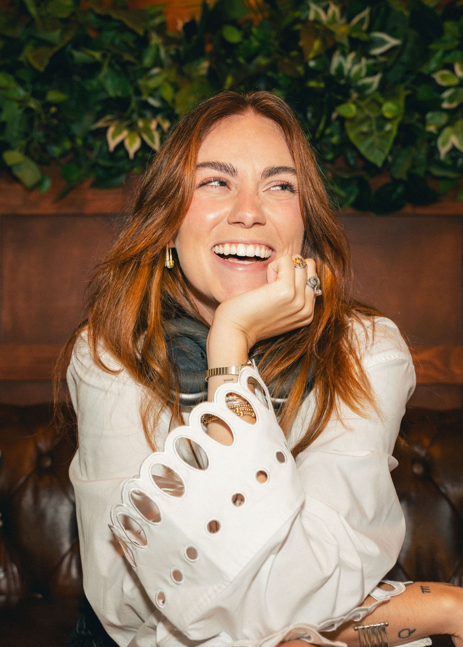 A laughing woman with auburn hair wearing a white cutout blouse sits in a cozy restaurant booth with green plants behind her.