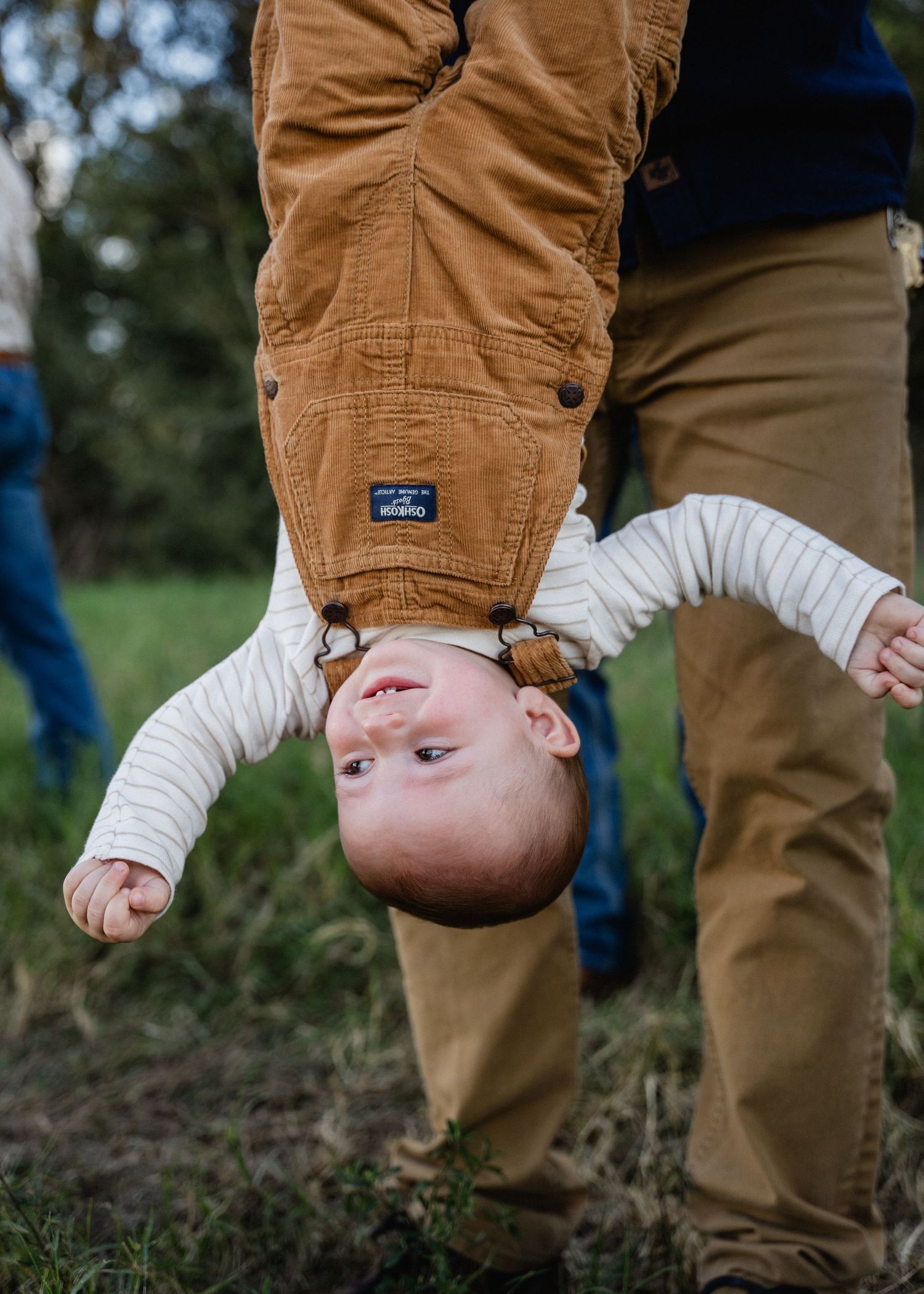 Father playfully holding toddler upside down by the legs outdoors, child smiling with joy.