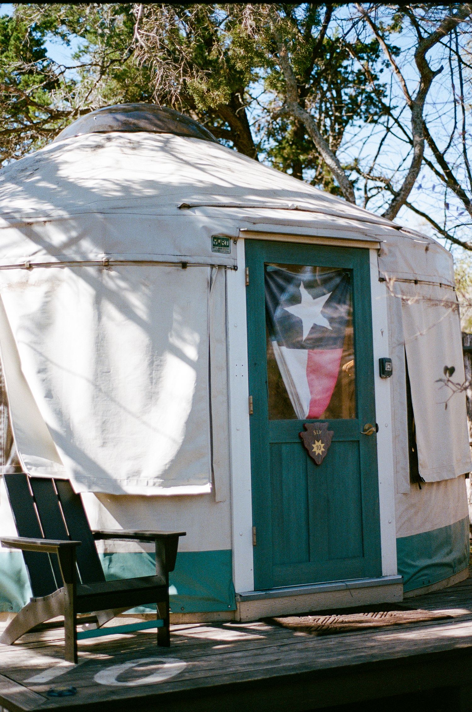 Rustic white yurt exterior with teal door featuring Texas flag, surrounded by trees with wooden deck chair.