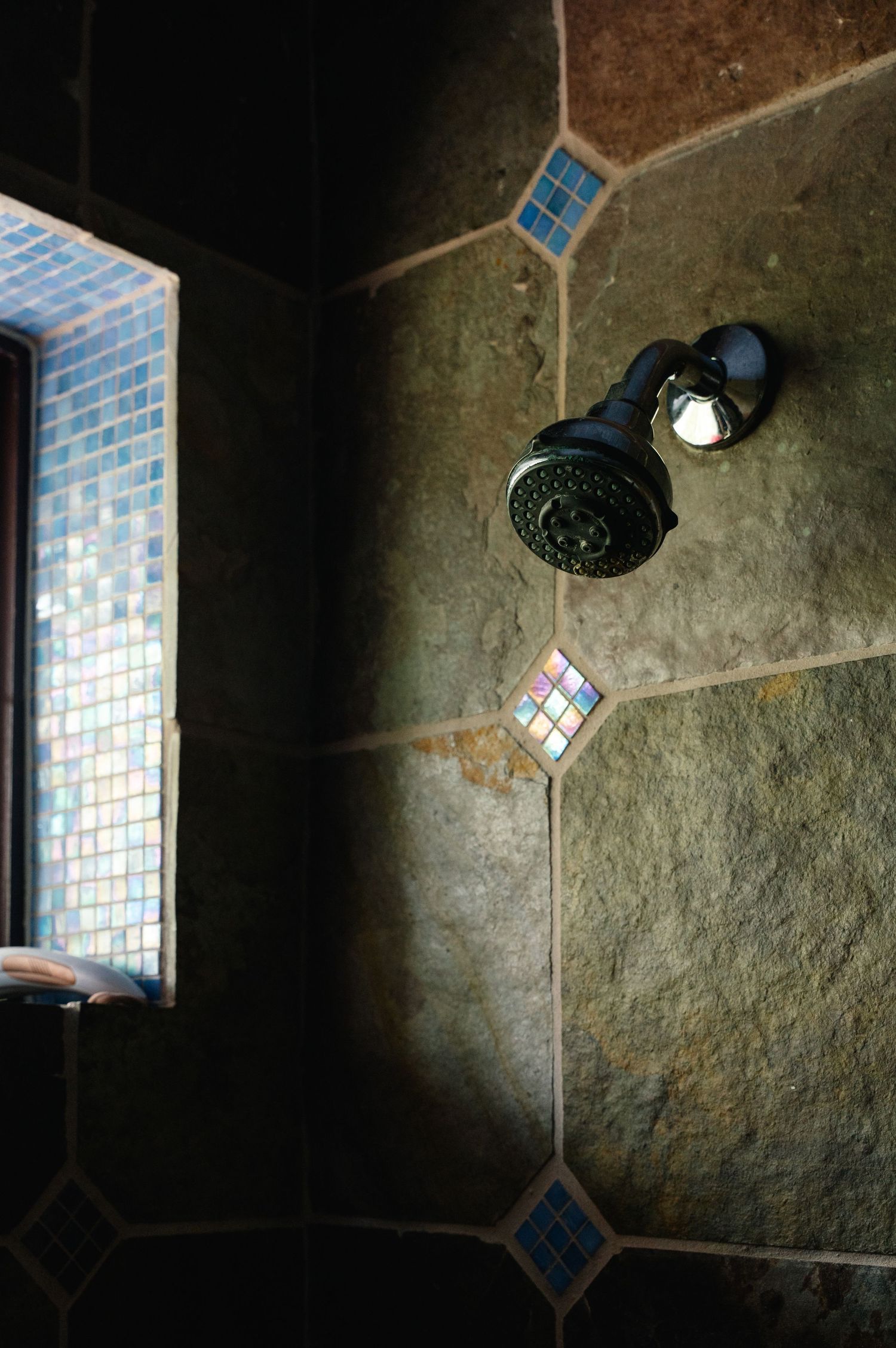 Stone tile shower wall with colorful mosaic accent tiles and dark metal showerhead beside a small window.