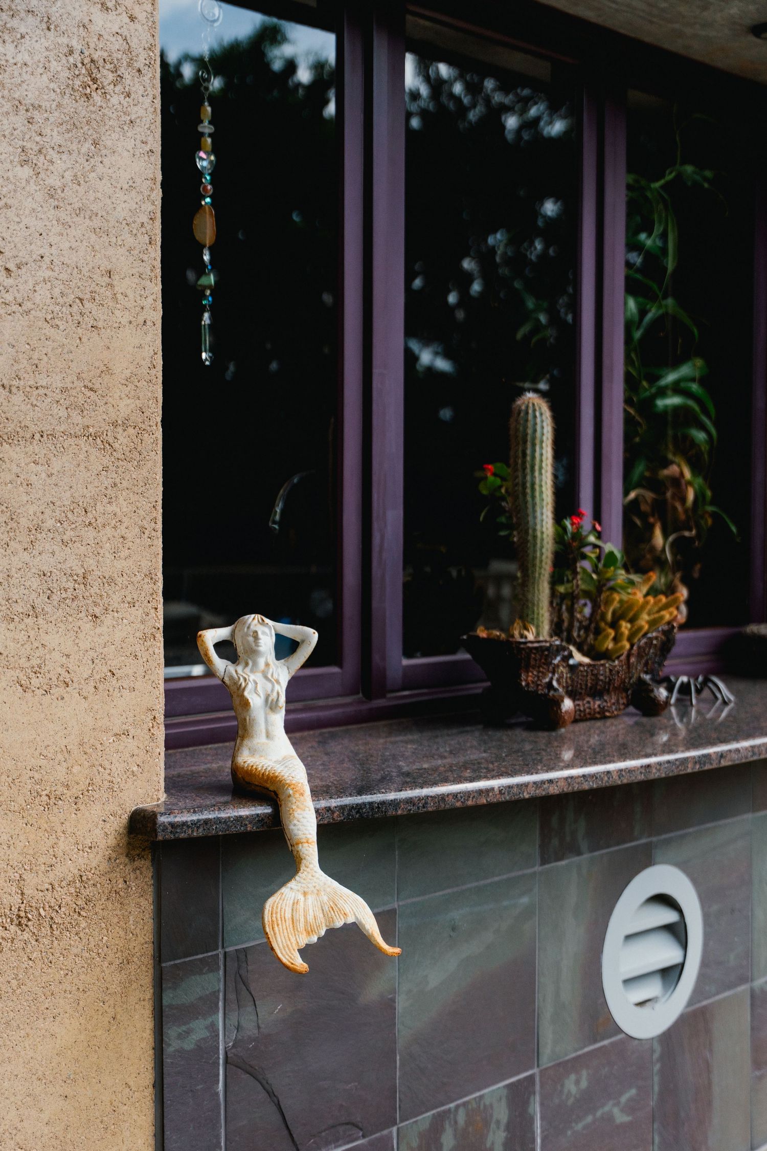 Decorative golden mermaid figurine perched on a granite windowsill beside potted cacti and succulents.