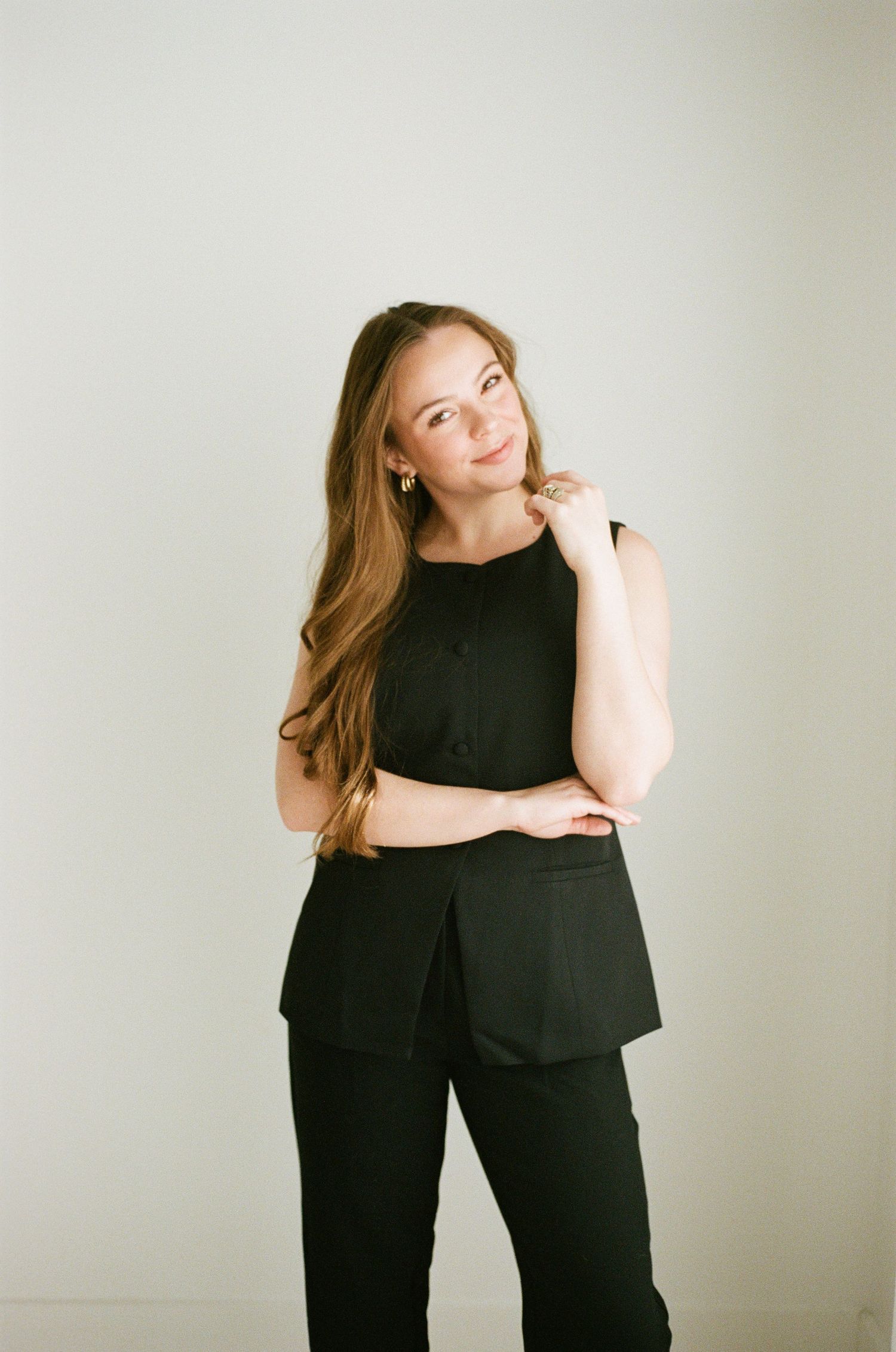 Young woman in all-black sleeveless top and trousers posing confidently against a light gray wall.