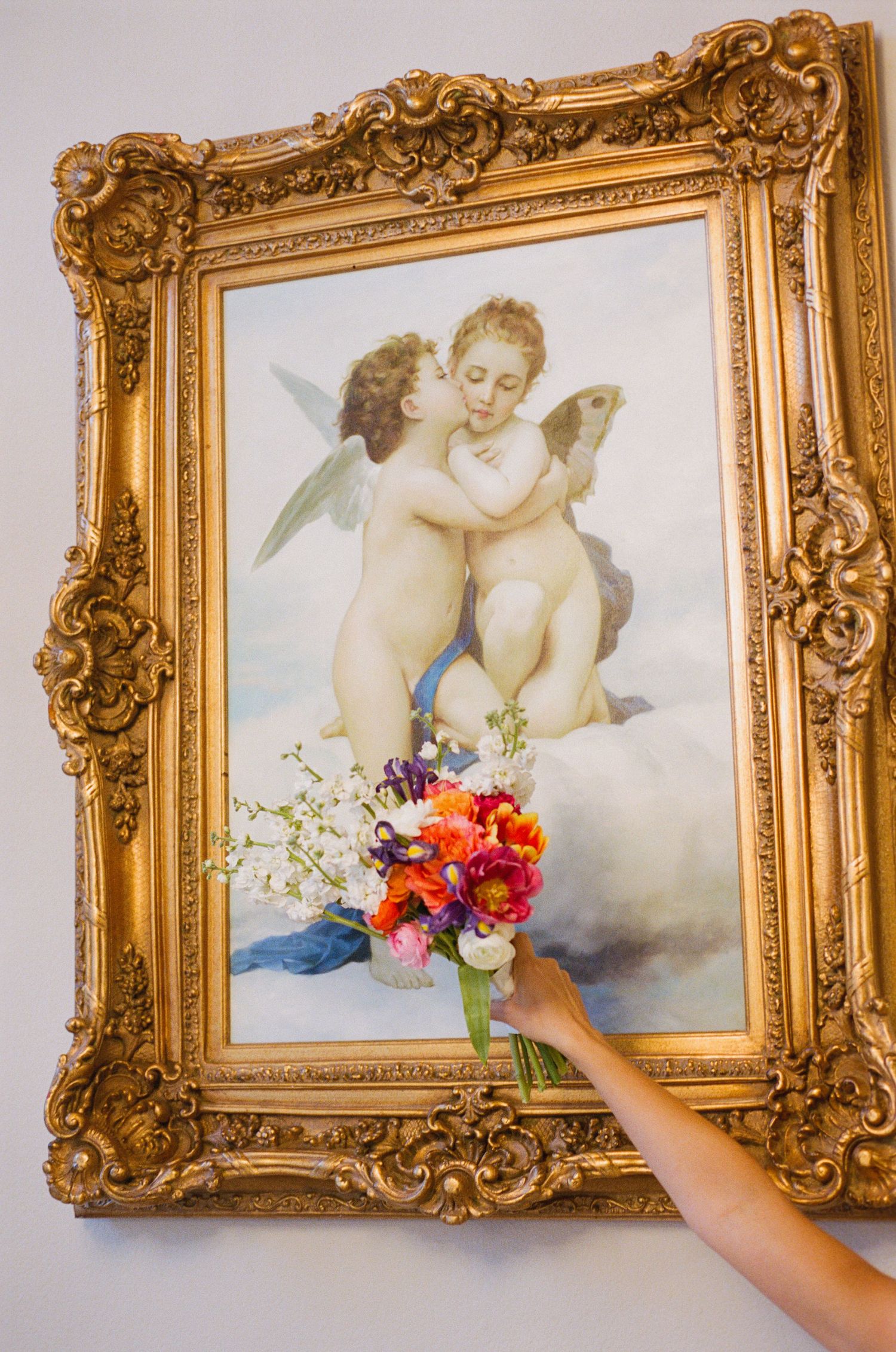 Hand holds vibrant colorful bridal bouquet in front of ornate gold-framed cherub painting on white wall.