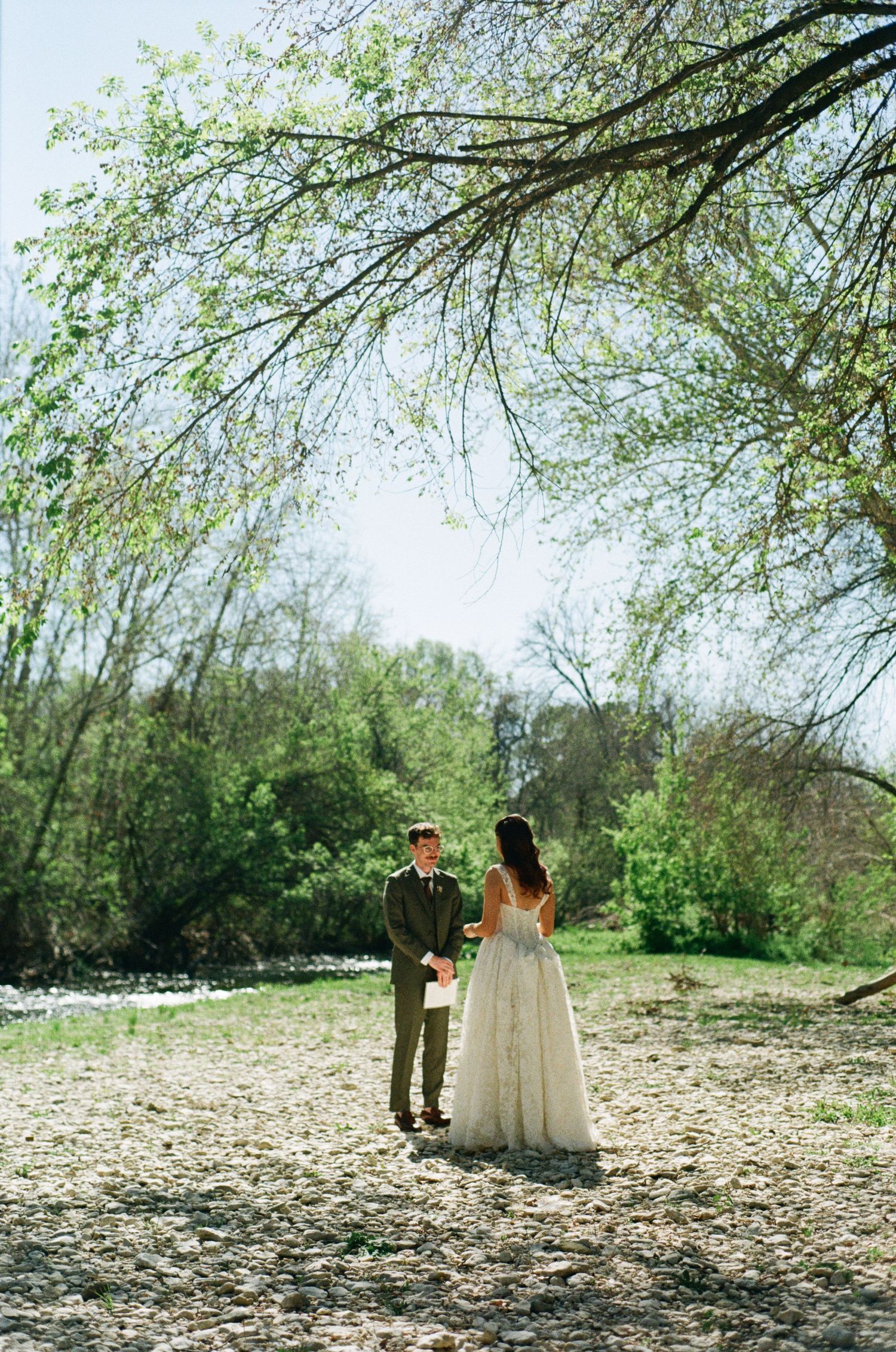 Bride and groom sharing first look moment on a rocky riverbank surrounded by lush spring trees.
