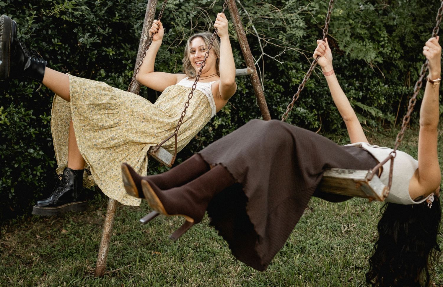 Two women in bohemian dresses laughing on wooden swings outdoors in a lush green garden setting.