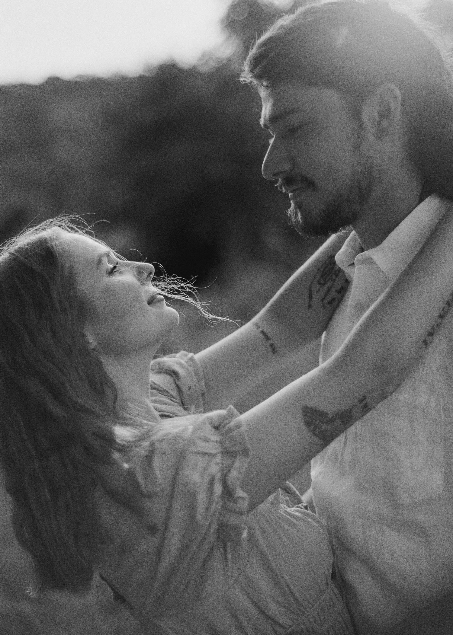 Black and white photo of a romantic couple embracing outdoors, woman gazing up at bearded tattooed man smiling.