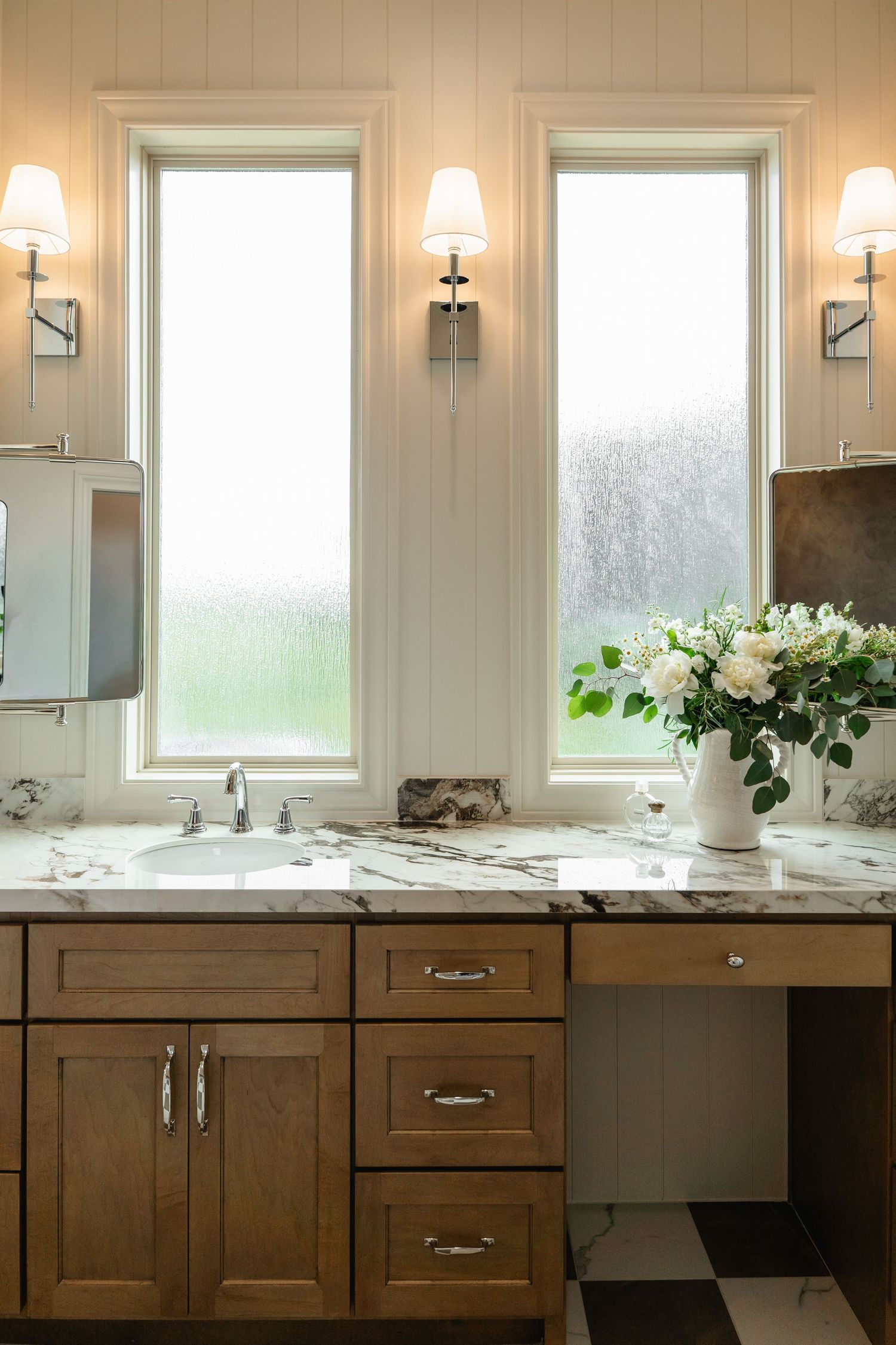 Elegant bathroom vanity with warm wood cabinets, marble countertop, white floral arrangement, and dual windows with sconce lighting.