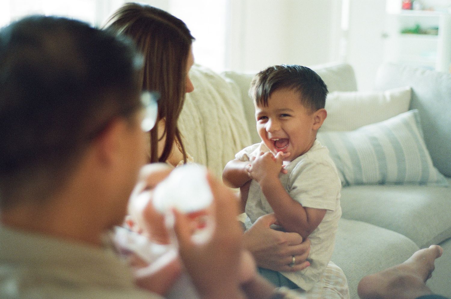 Happy toddler boy laughing joyfully while sitting with mother and newborn sibling on a white sofa.