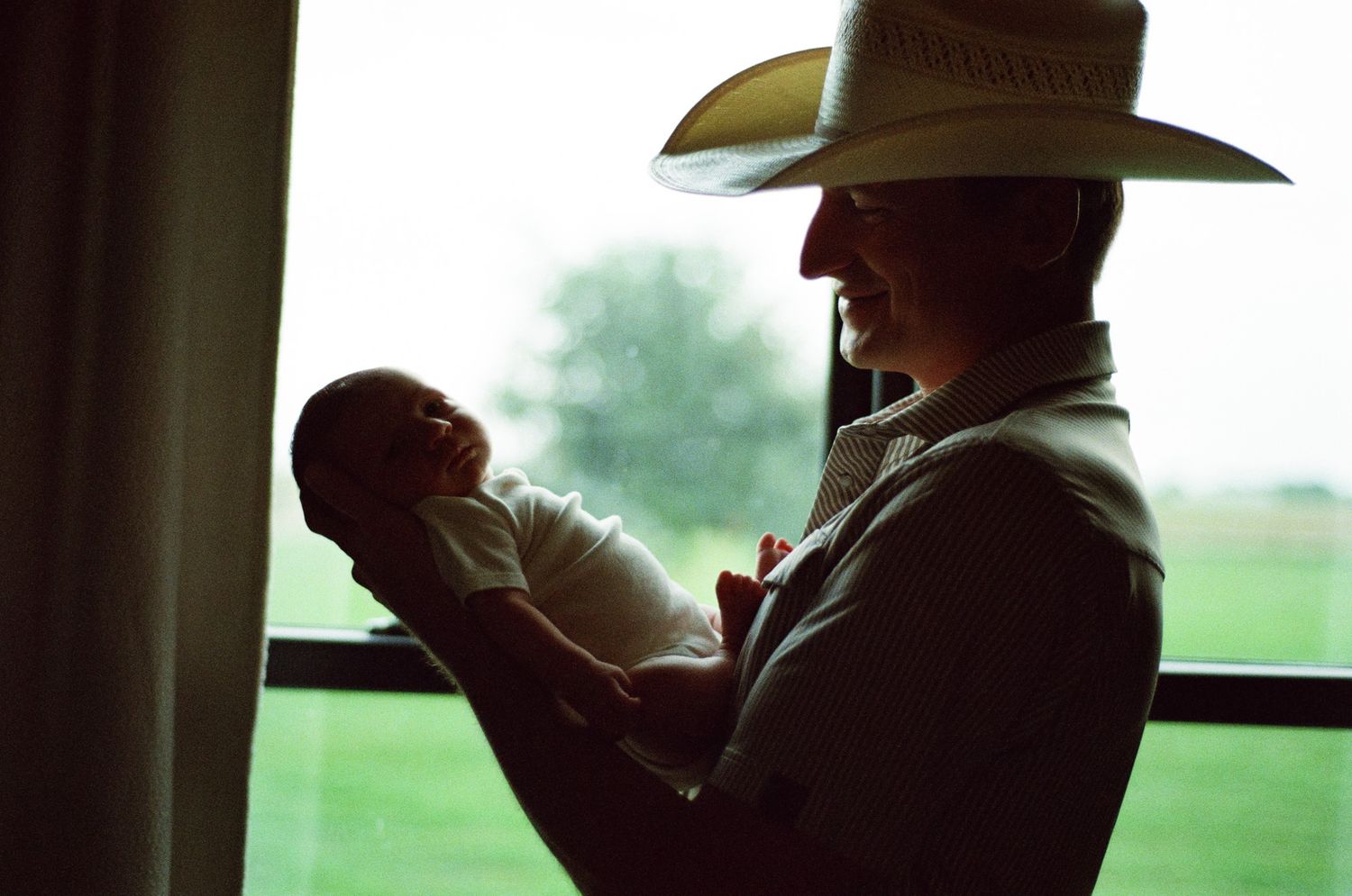 Silhouette of a man in a cowboy hat cradling a newborn baby by a bright window on a farm.