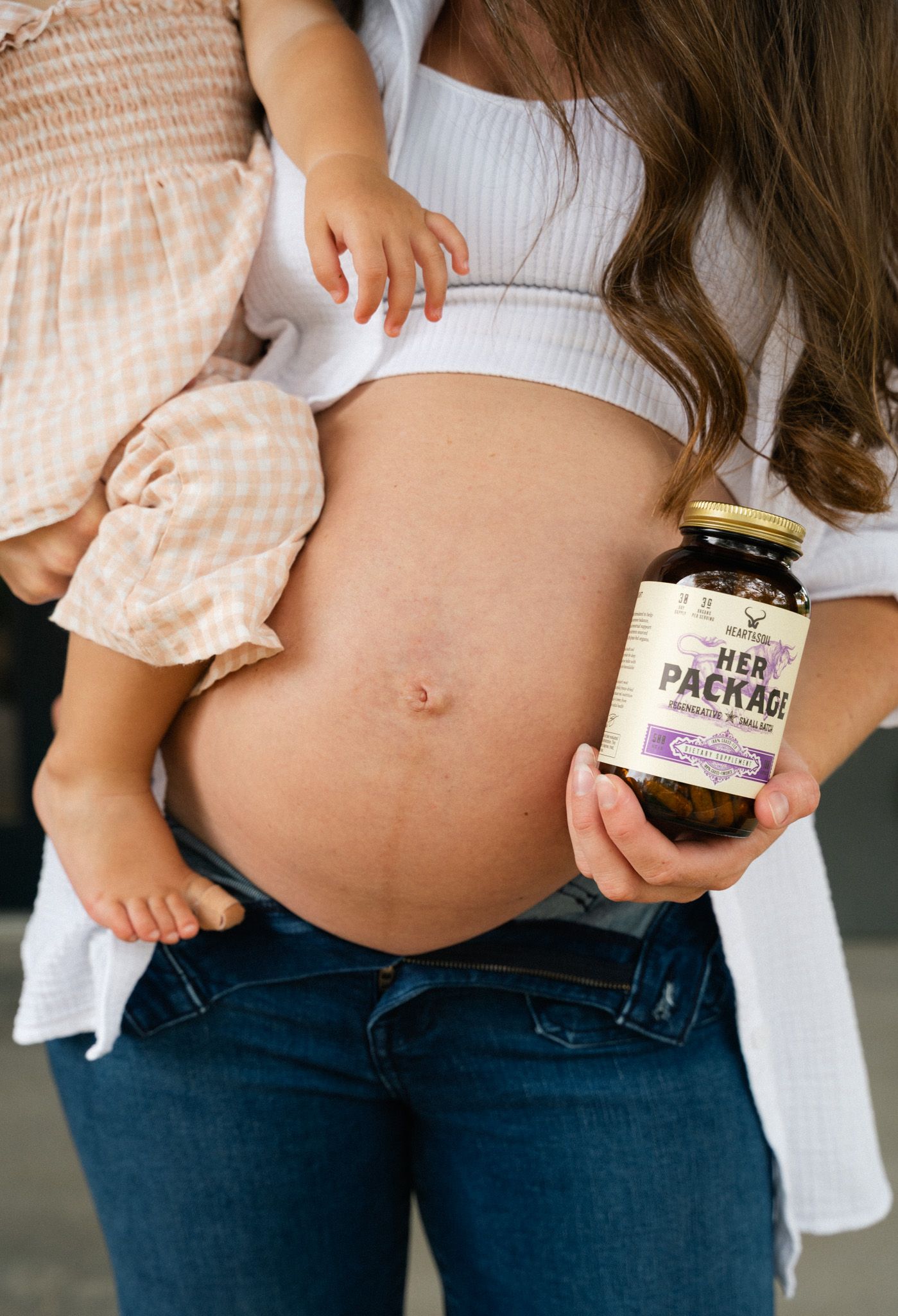 Pregnant woman holding a baby and a jar of Her Package prenatal supplements, showcasing maternal wellness products.