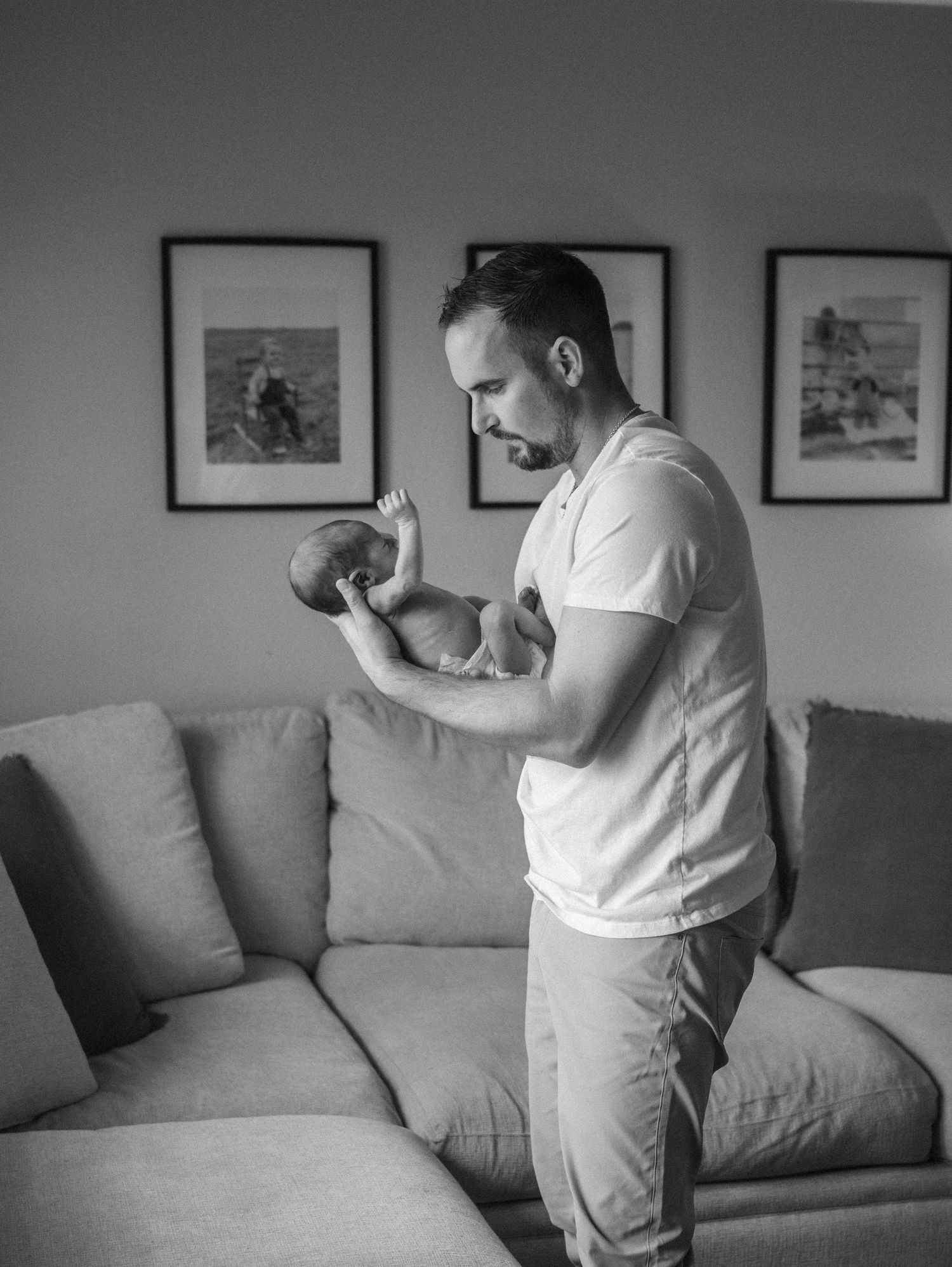 Black and white photo of father holding newborn baby while standing near gray sofa at home.