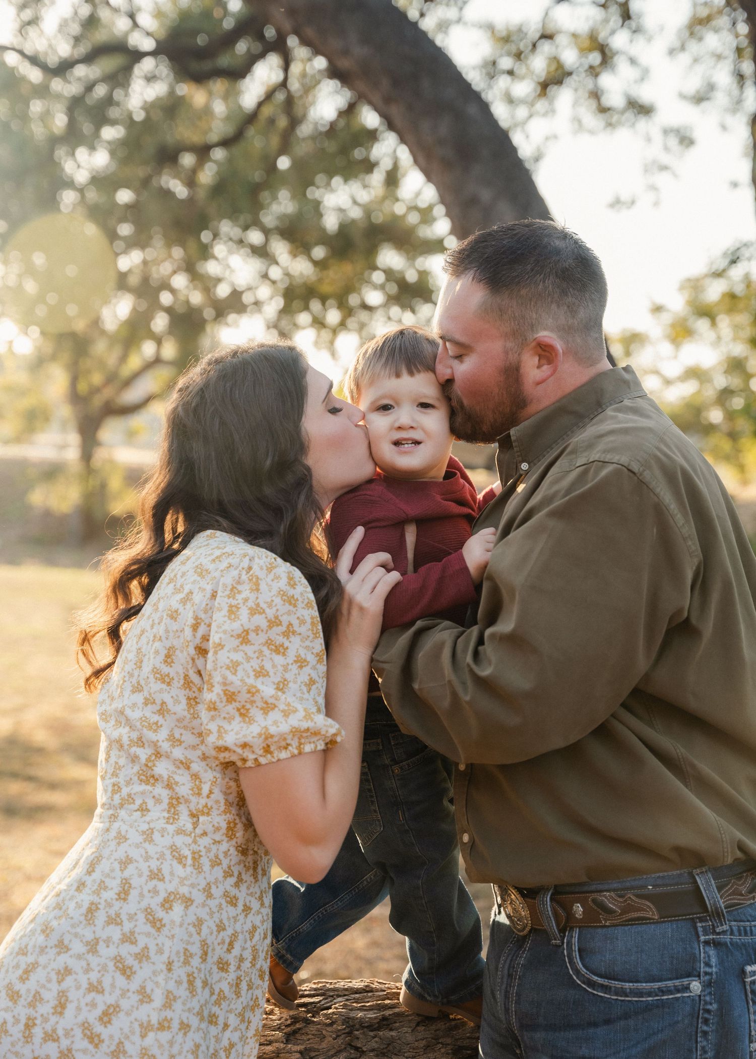Two parents lovingly kiss their toddler son on the cheeks during a warm golden hour family photo session.