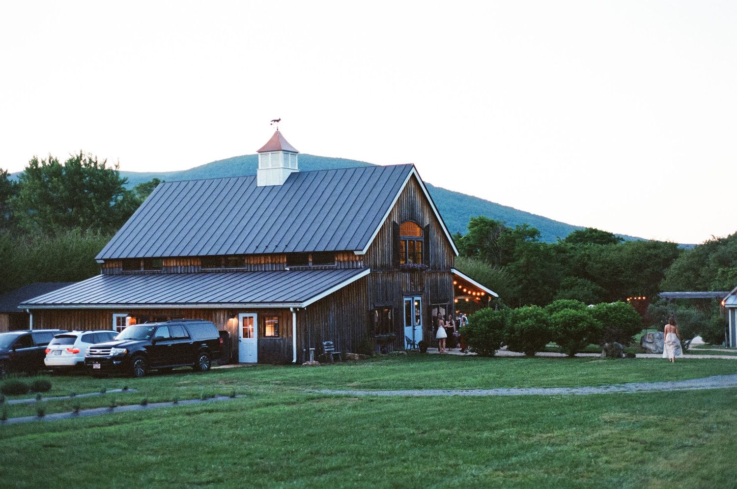 Charming rustic barn venue with metal roof, cupola, and mountain backdrop at dusk.