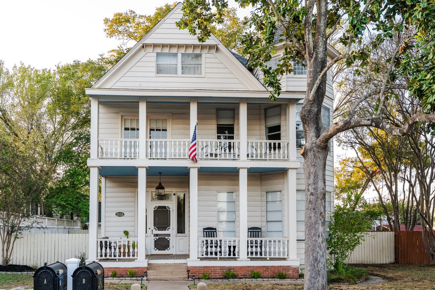 Victorian-style two-story home with white siding, wrap-around porches, American flag, and mature trees in front yard.