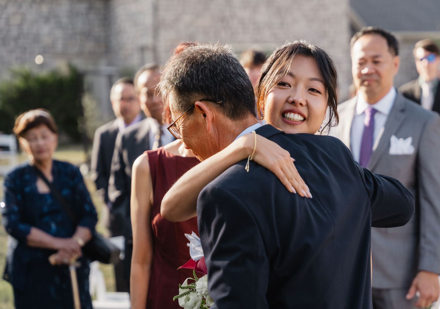 Bride shares joyful hug with family member during outdoor wedding ceremony with guests nearby.