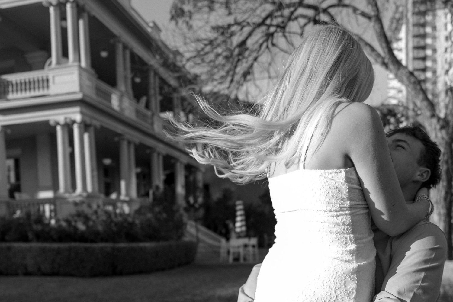 Black and white photo of a couple embracing outdoors near a historic Victorian house, woman's blonde hair flowing.