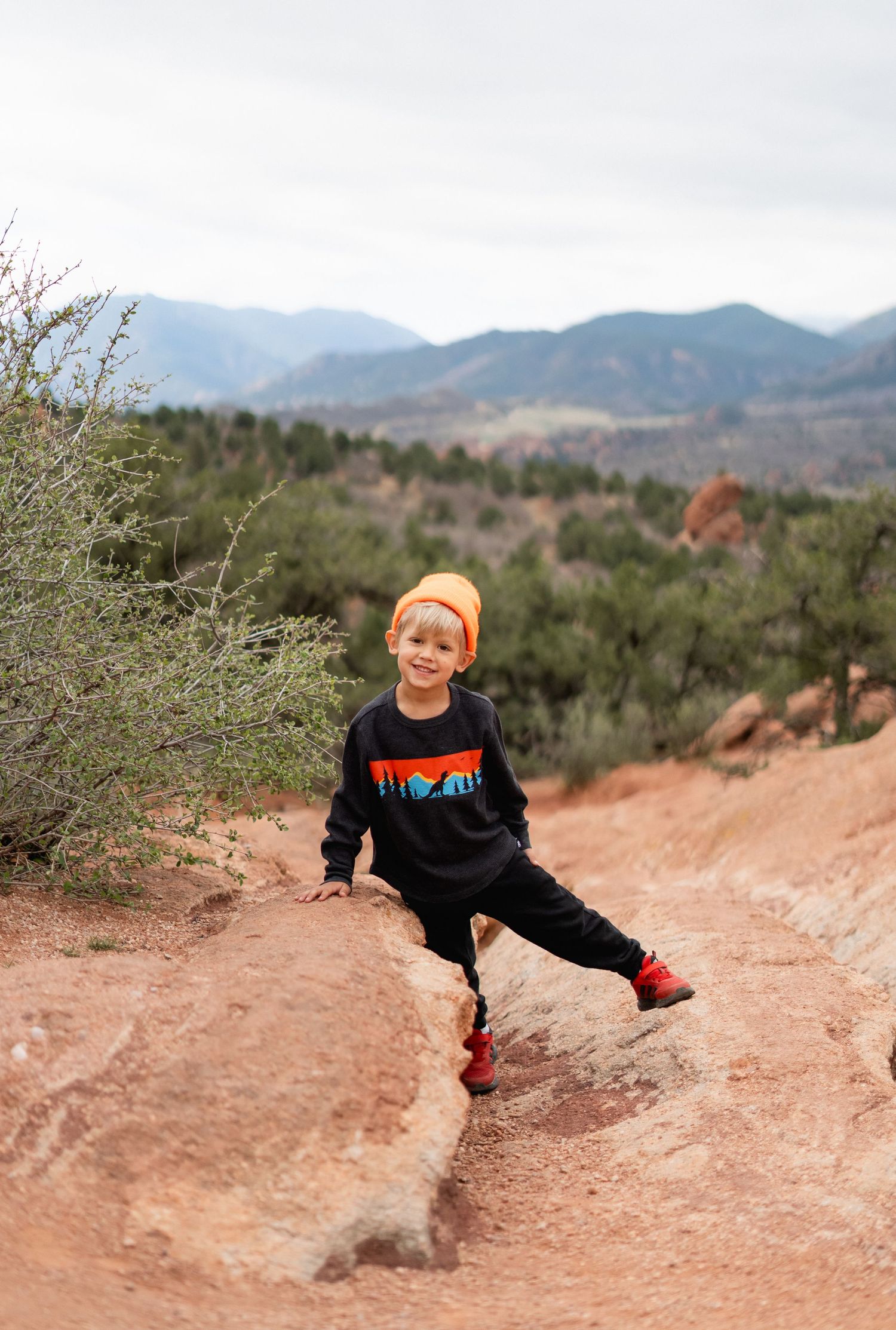 Young boy in orange beanie and black sweatshirt poses on red rock trail at Garden of the Gods, Colorado.