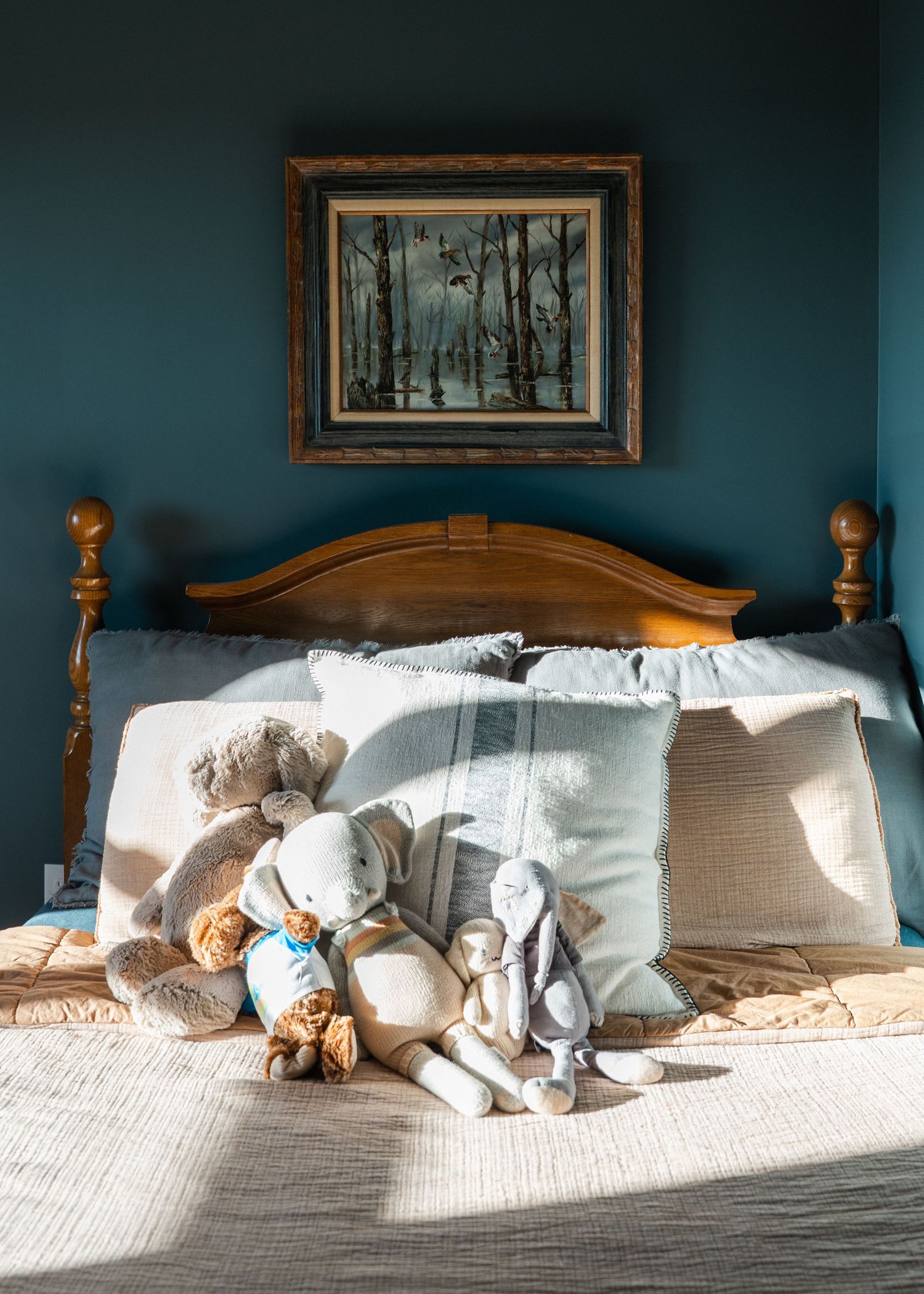 Cozy bedroom with teal accent wall, wooden headboard, stuffed animal toys, and a landscape painting above the bed.