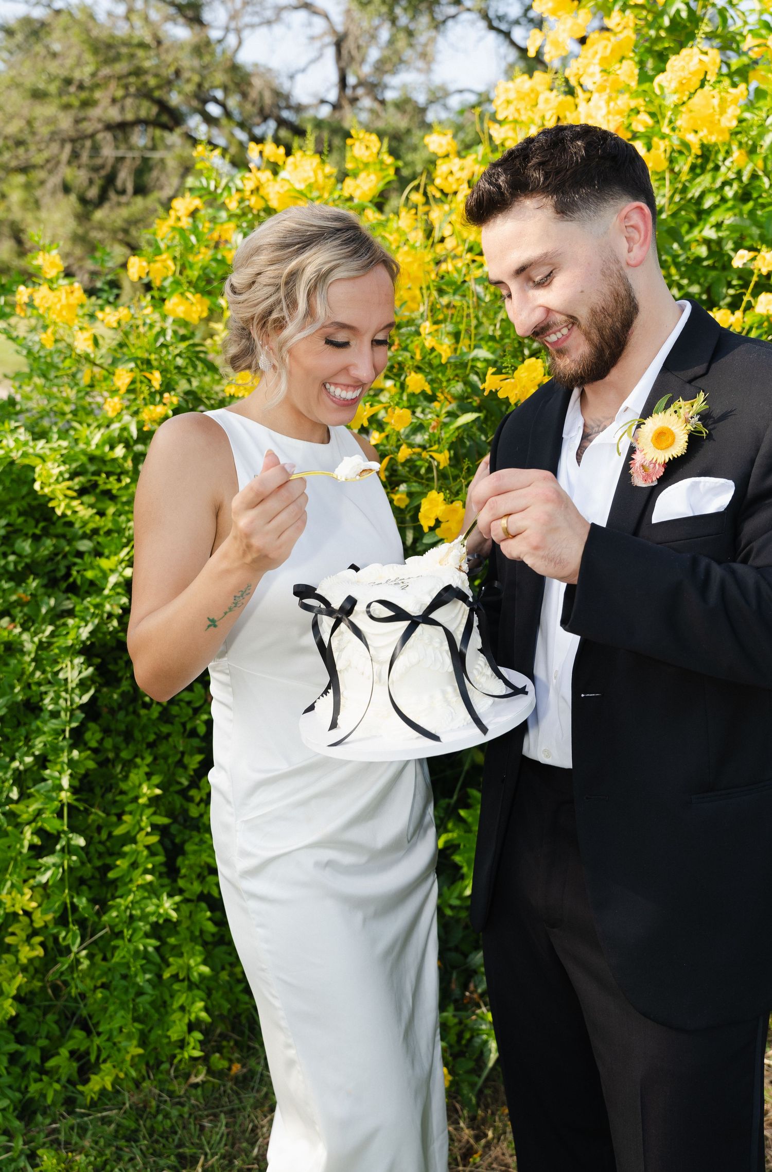 Bride and groom smiling while cutting small wedding cake in front of yellow flowering bushes.
