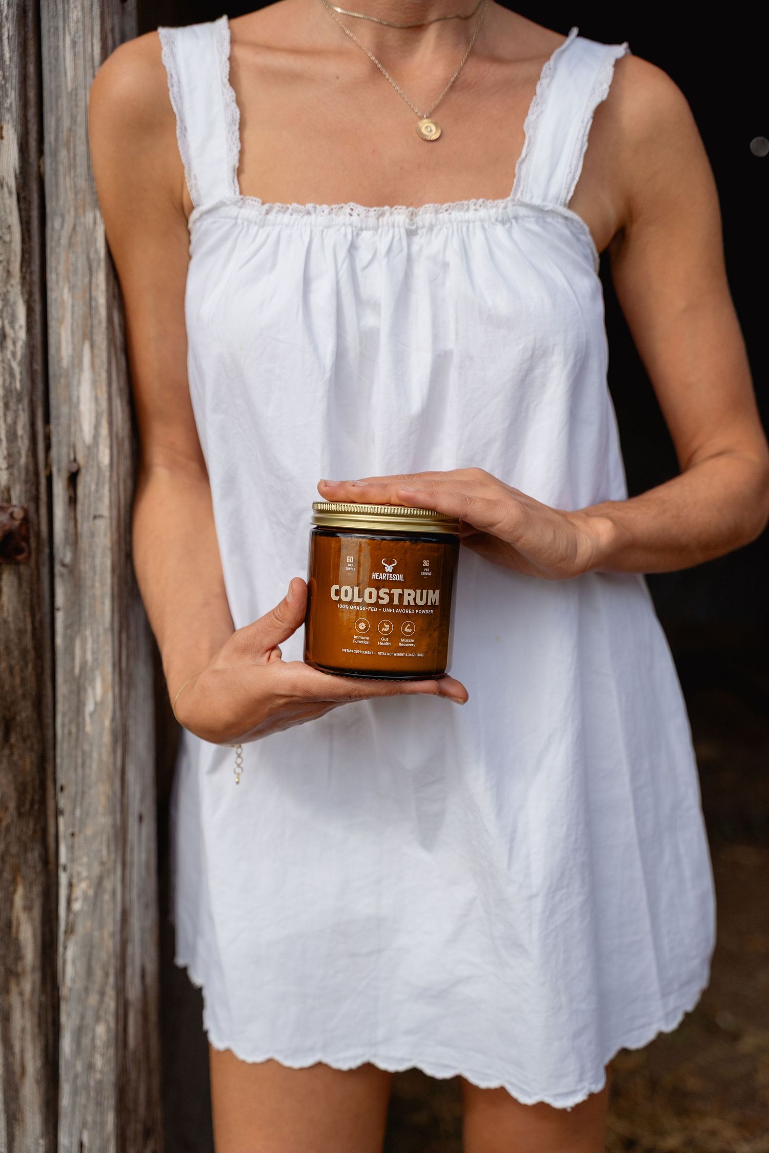Woman in white sundress holding a jar of Colostrum supplement near a barn.