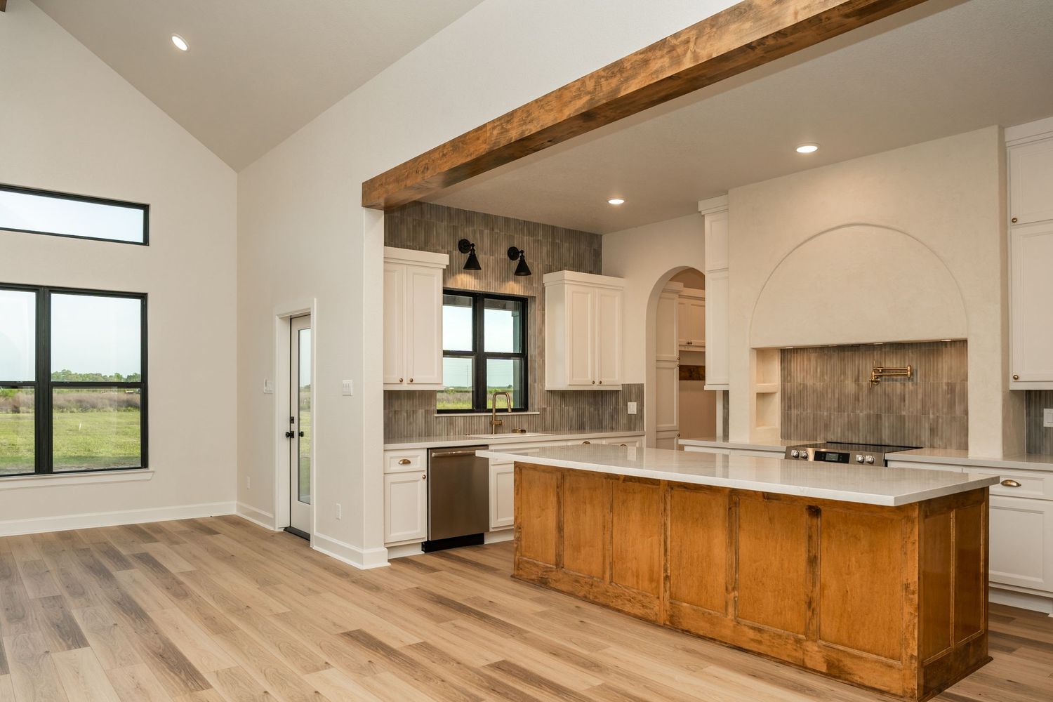 Open kitchen with high vaulted ceilings, wood beam, white cabinets, wood island, and arched backsplash accent.