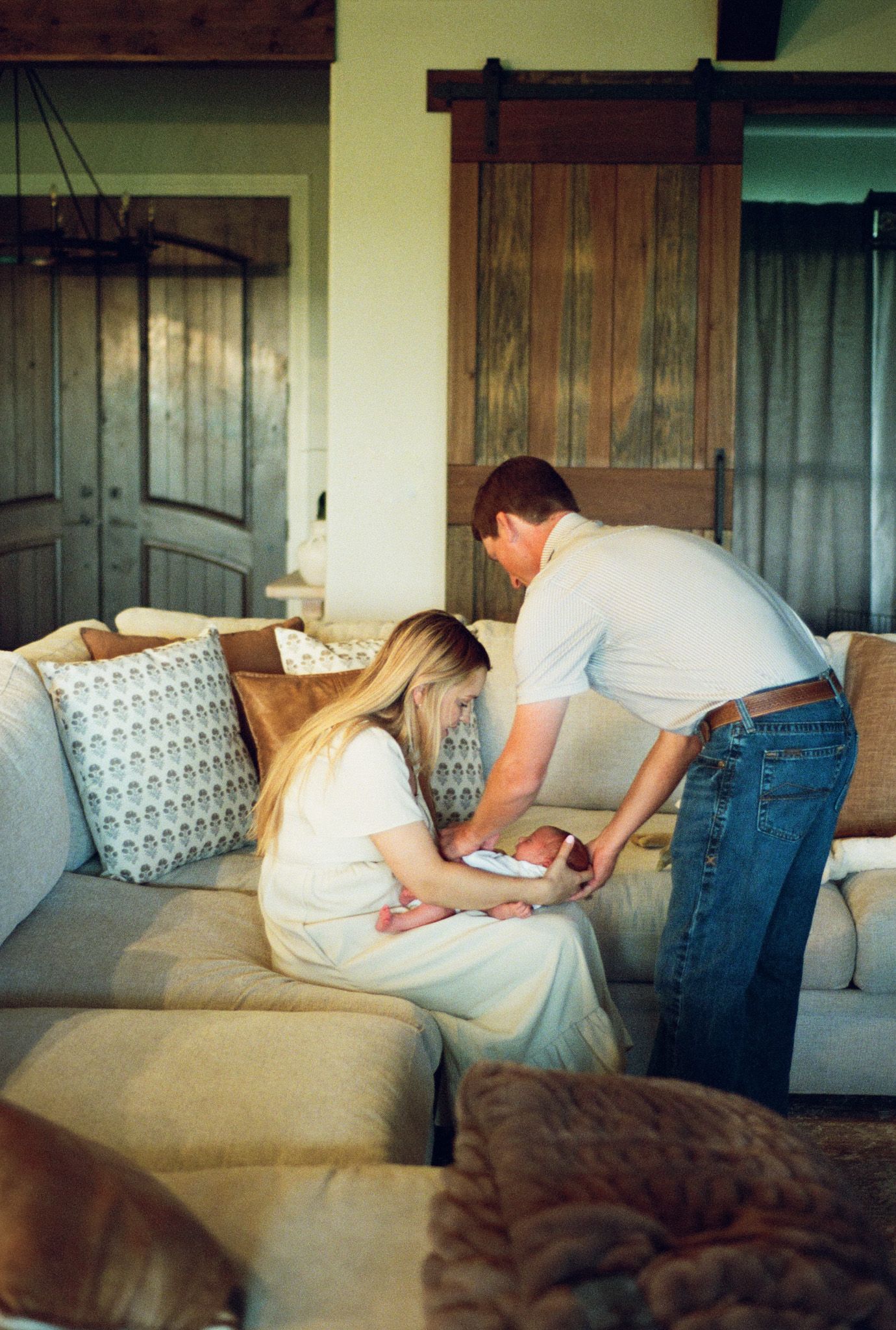 New parents leaning over couch together carefully tending to their newborn baby in rustic living room.