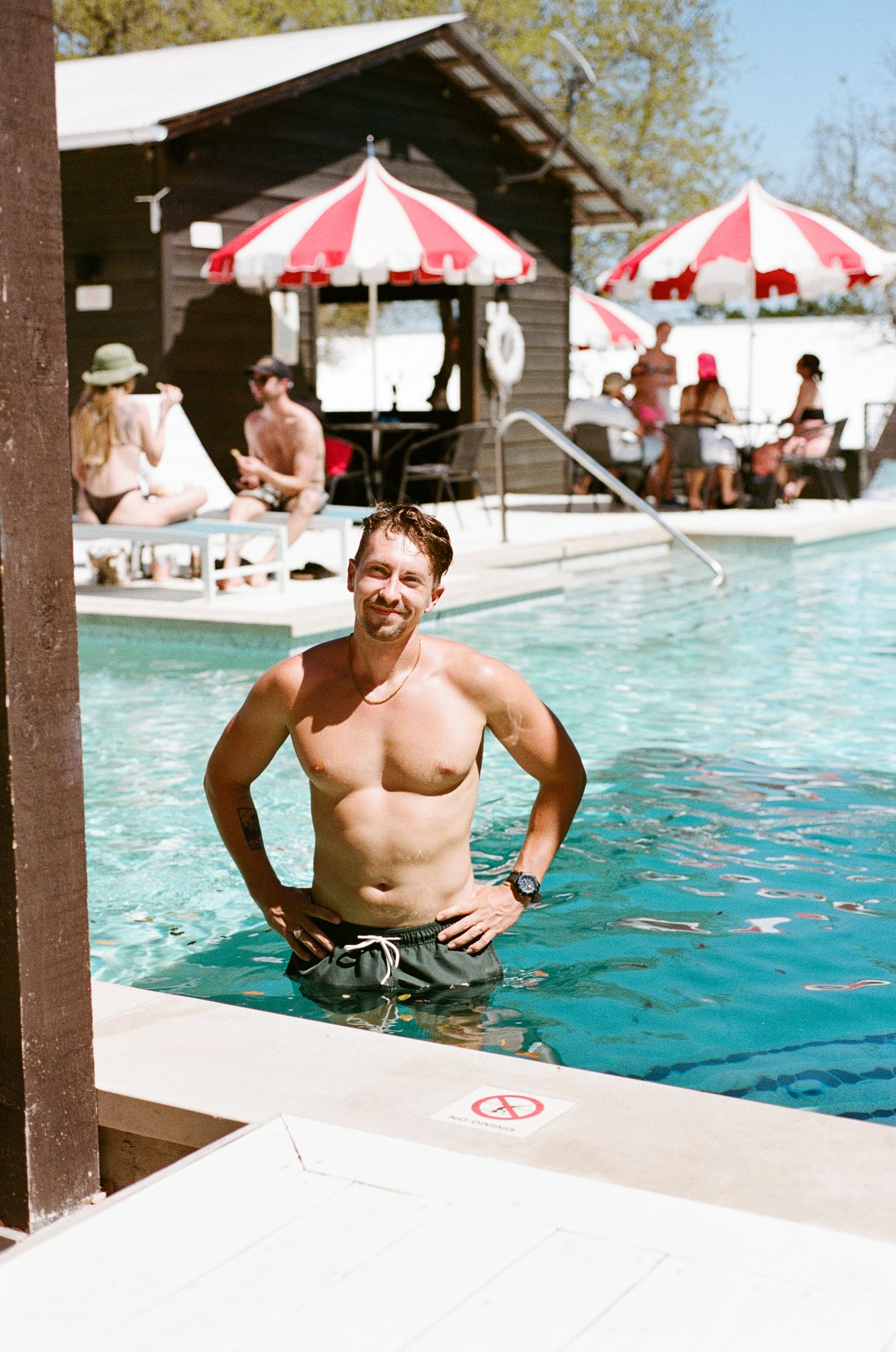 Smiling man standing in a resort pool with red and white striped umbrellas and guests in the background.