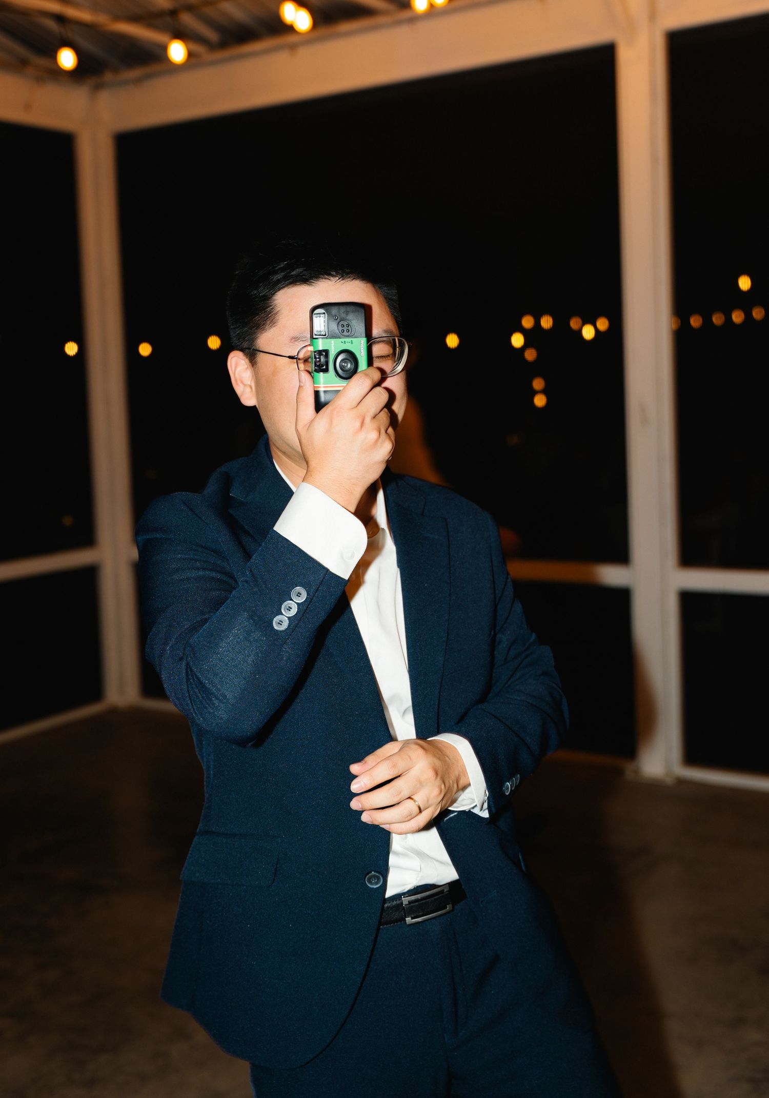Dapper wedding guest in navy suit photographs event using a vintage-style camera at night.