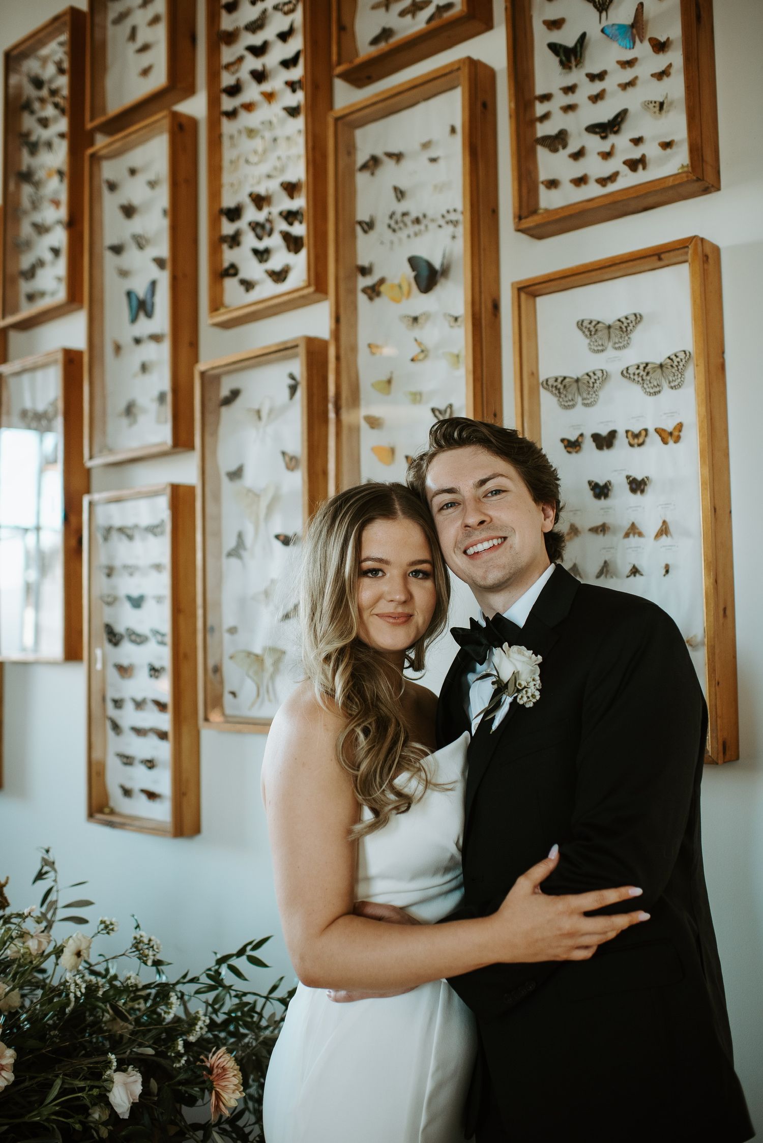 Smiling newlywed couple embracing in front of framed butterfly collection wall display.