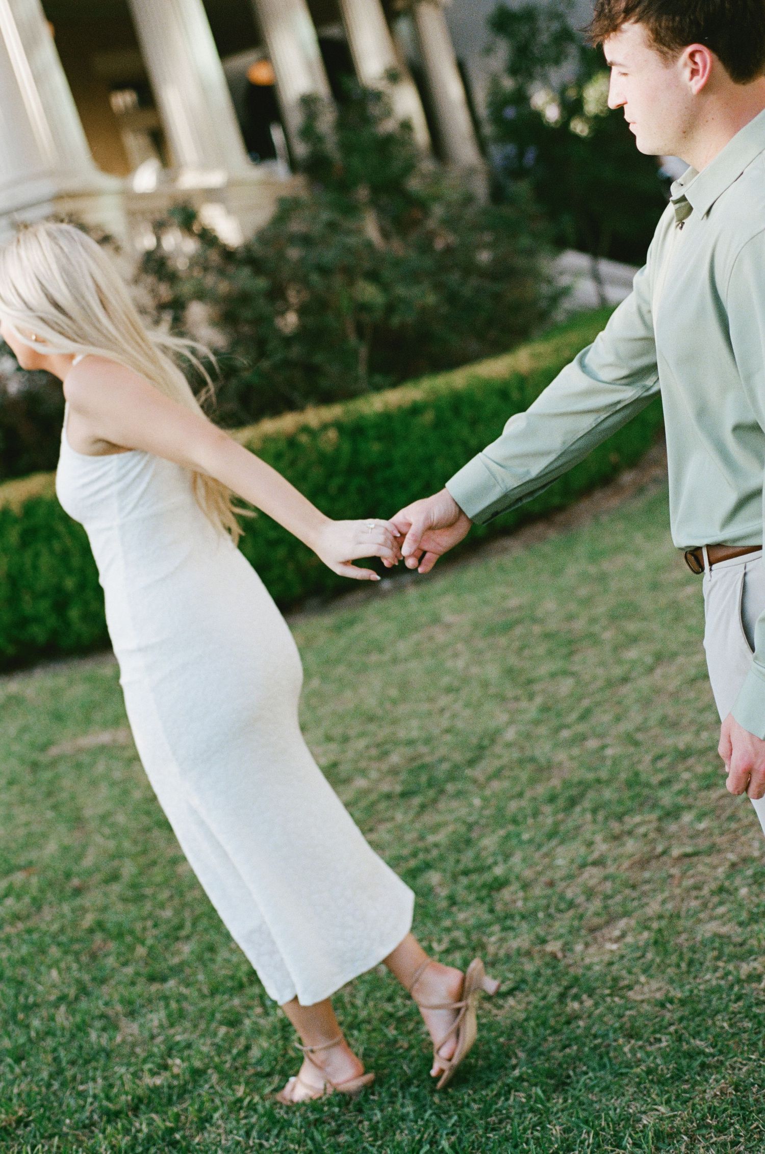 A couple holds hands on a green lawn, the woman in a white midi dress and the man in a sage green shirt.