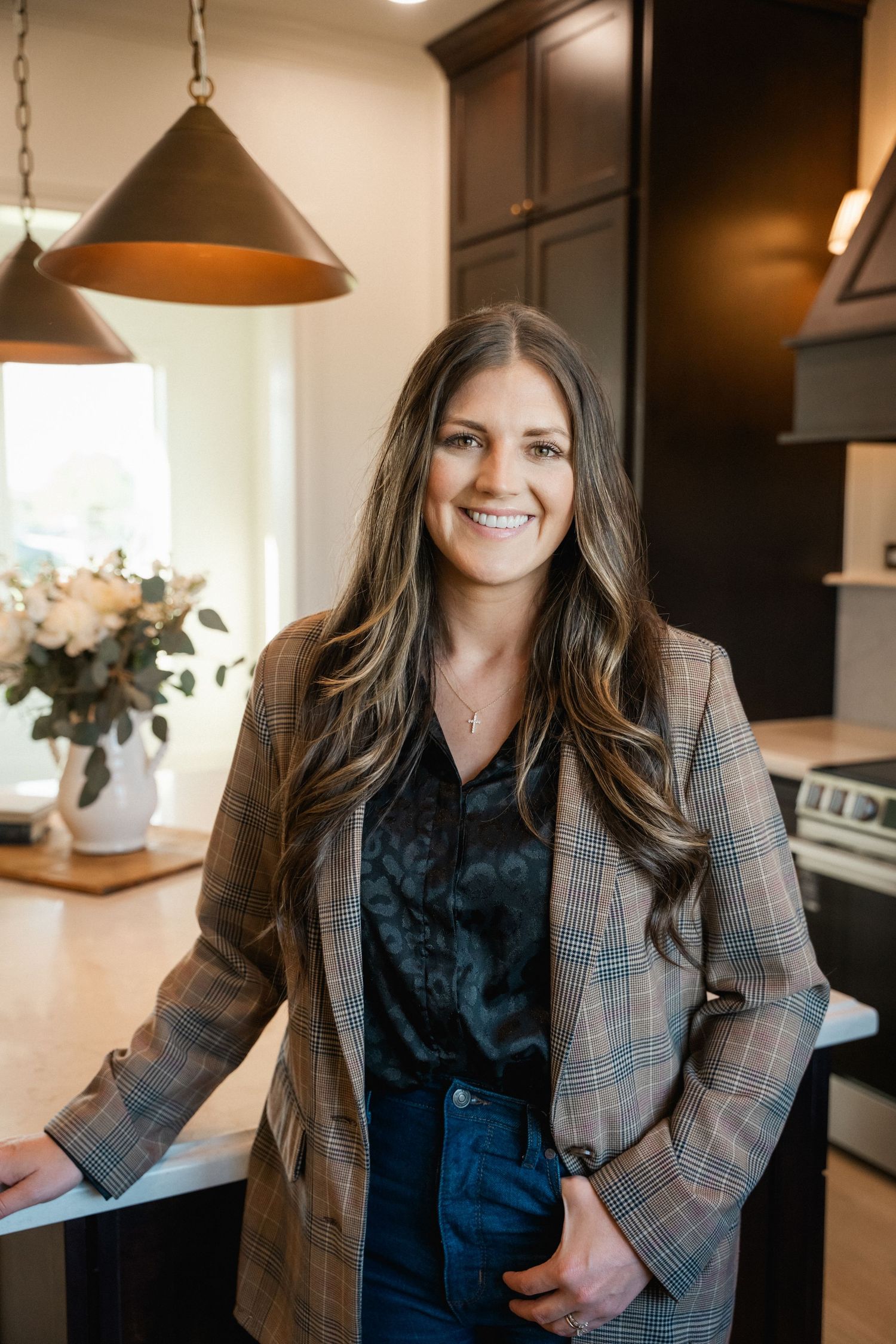 Smiling woman with long brown hair wearing a plaid blazer leans against a kitchen counter in a modern home.