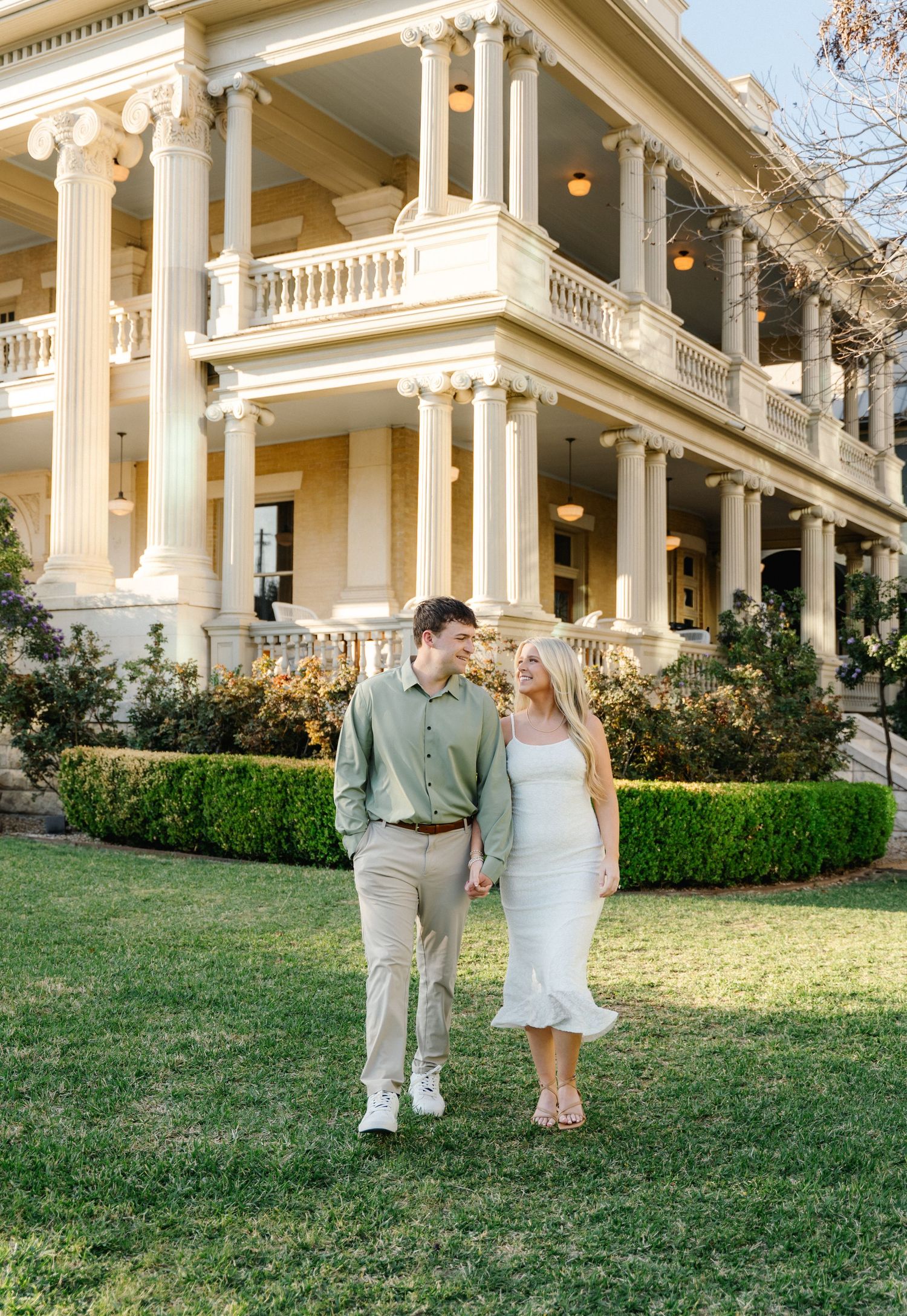 Engaged couple holding hands walking on green lawn before grand white columned mansion at golden hour.