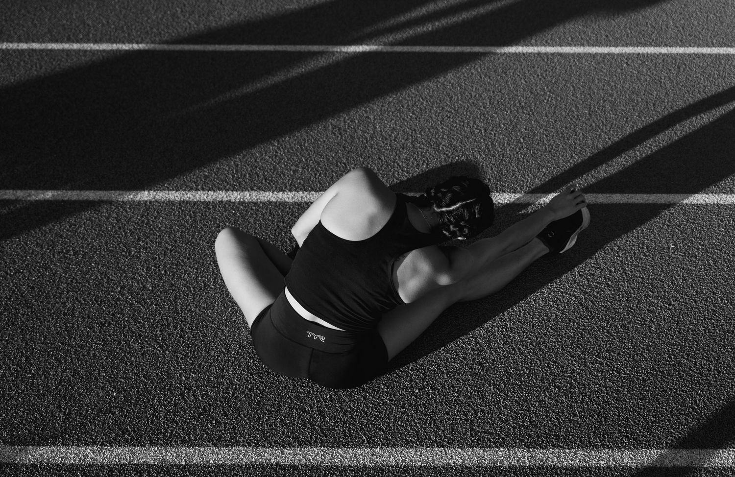 Black and white photo of an athlete stretching on a running track, seated with legs extended in a flexibility exercise.