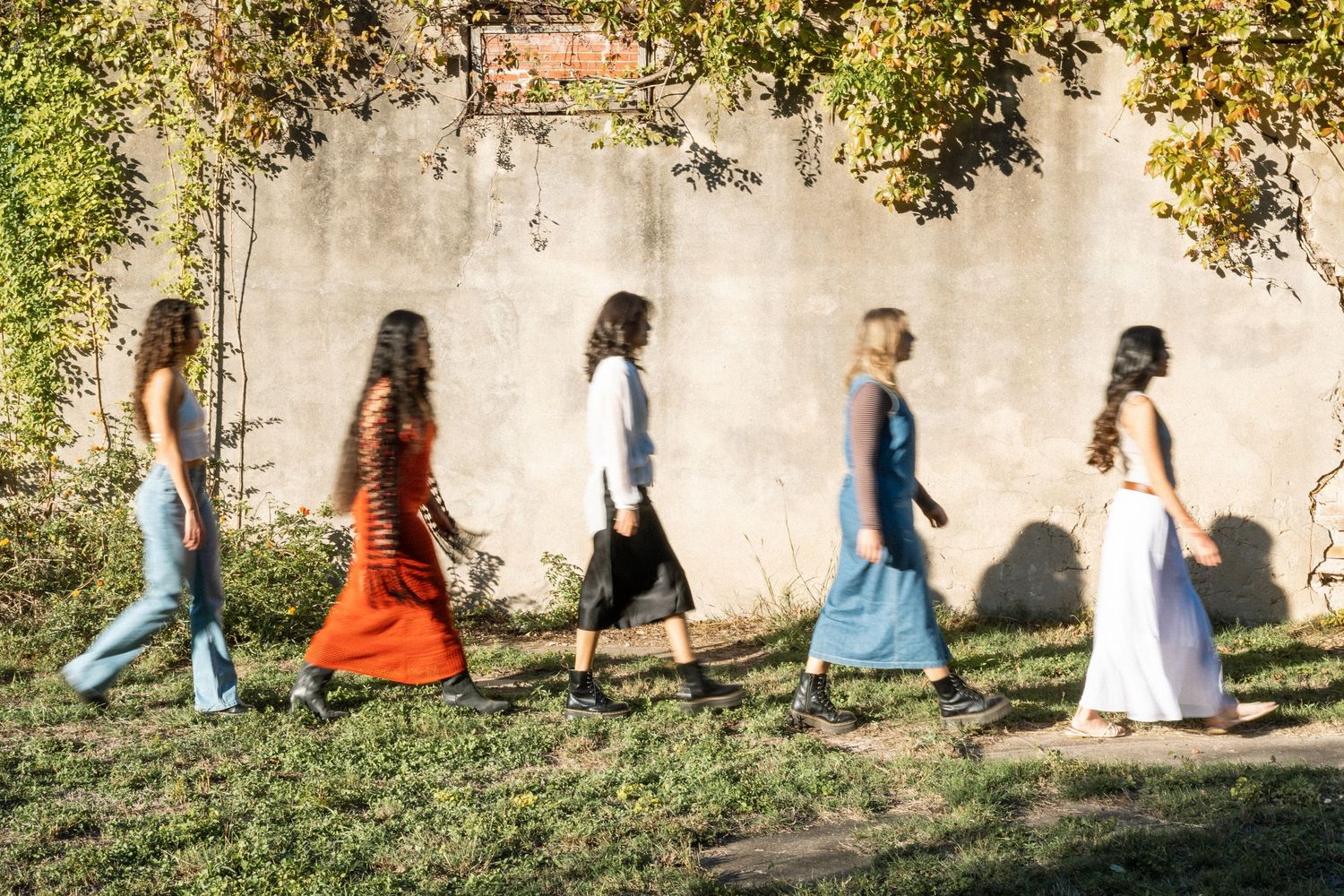 Four women in colorful dresses walking outdoors along a sunlit grass path beside a concrete wall.